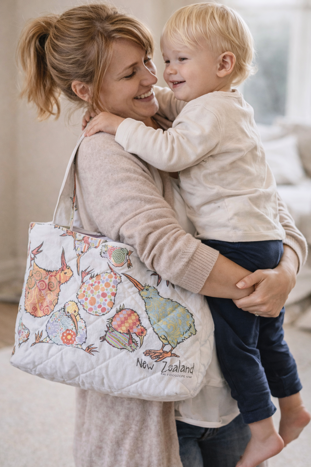 A woman holding a young blonde boy in her arms, smiling and hugging tightly, with a New Zealand-themed bag over her shoulder.