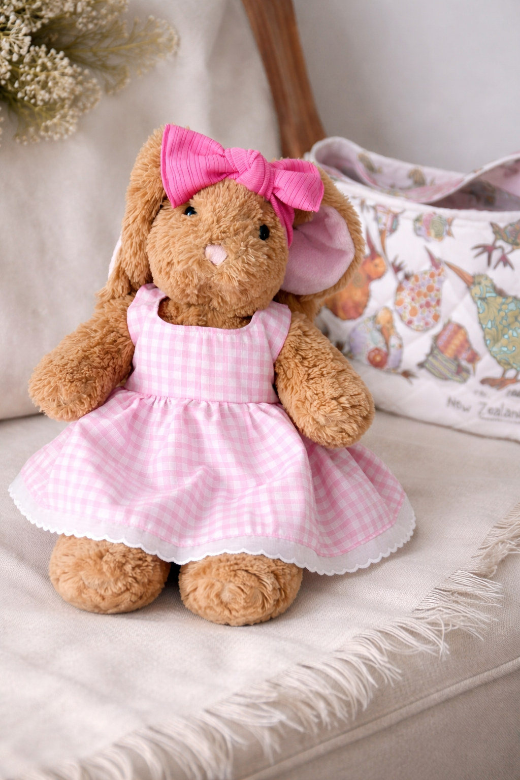 A plush toy bunny dressed in a pink checkered dress with scalloped edges, a matching pink bow on its head, and pink lining inside its ears, sitting on a beige surface with a pillow that features a map of New Zealand and dried flowers in the background.