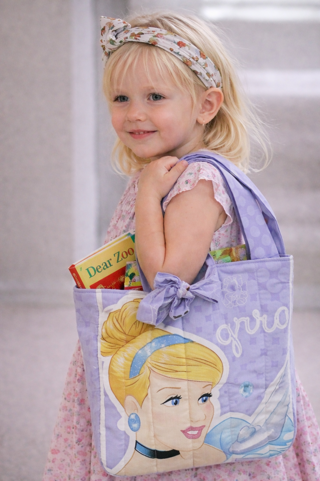 A young girl with blonde hair wearing a floral headband, carrying a purple Disney princess-themed bag filled with books, including a "Dear Zoo" book, and wearing a pink floral dress.