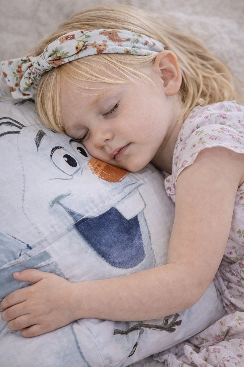 A young girl with blonde hair, wearing a floral headband and pink floral pajamas, sleeping peacefully on a pillow with a cartoon snowman design.