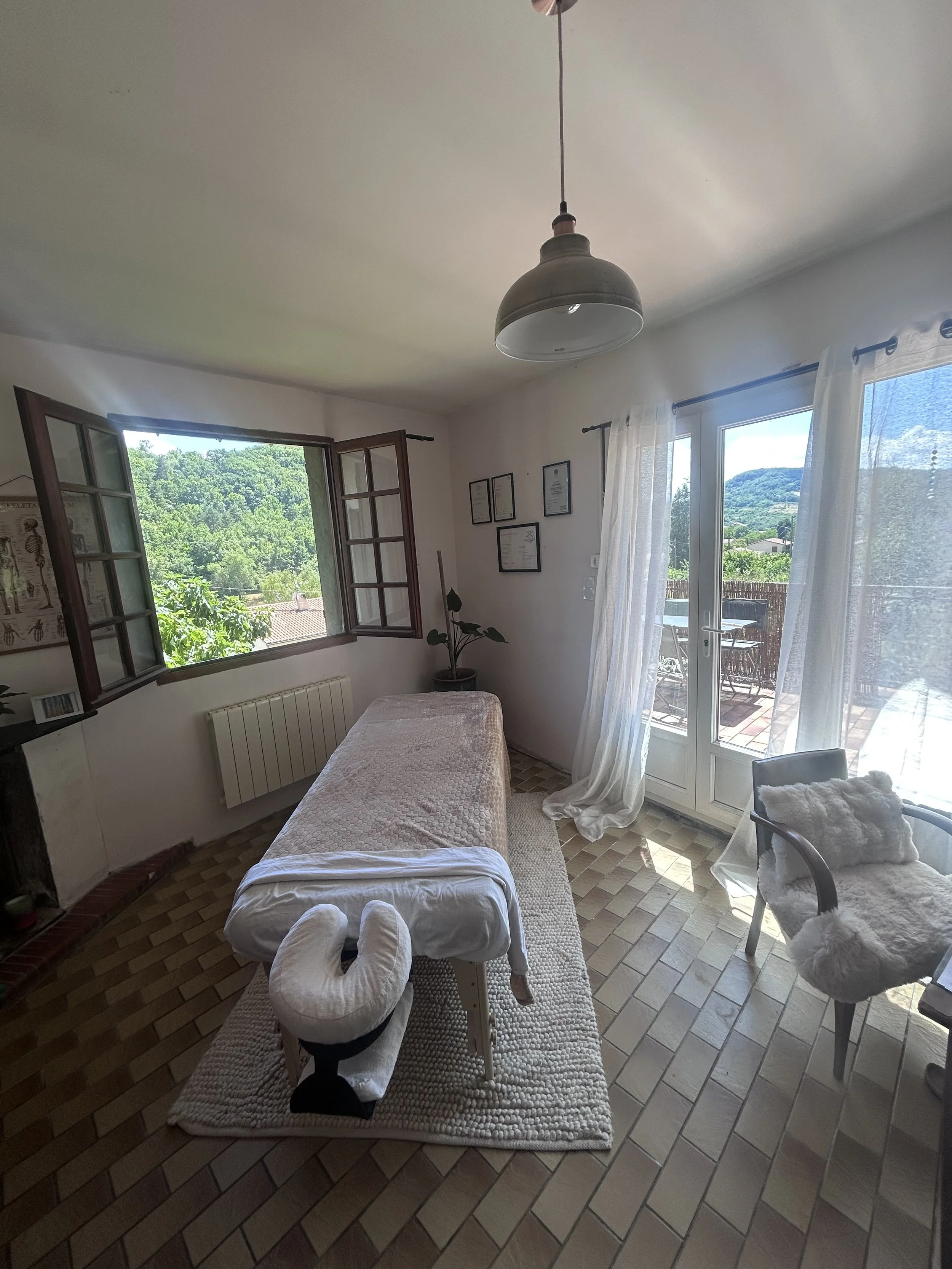 Une pièce lumineuse avec une table de massage, une fenêtre ouverte laissant entrer la lumière naturelle, et un décor intérieur simple avec un fauteuil, des cadres sur le mur et une porte donnant sur un balcon avec vue sur des montagnes et des arbres.