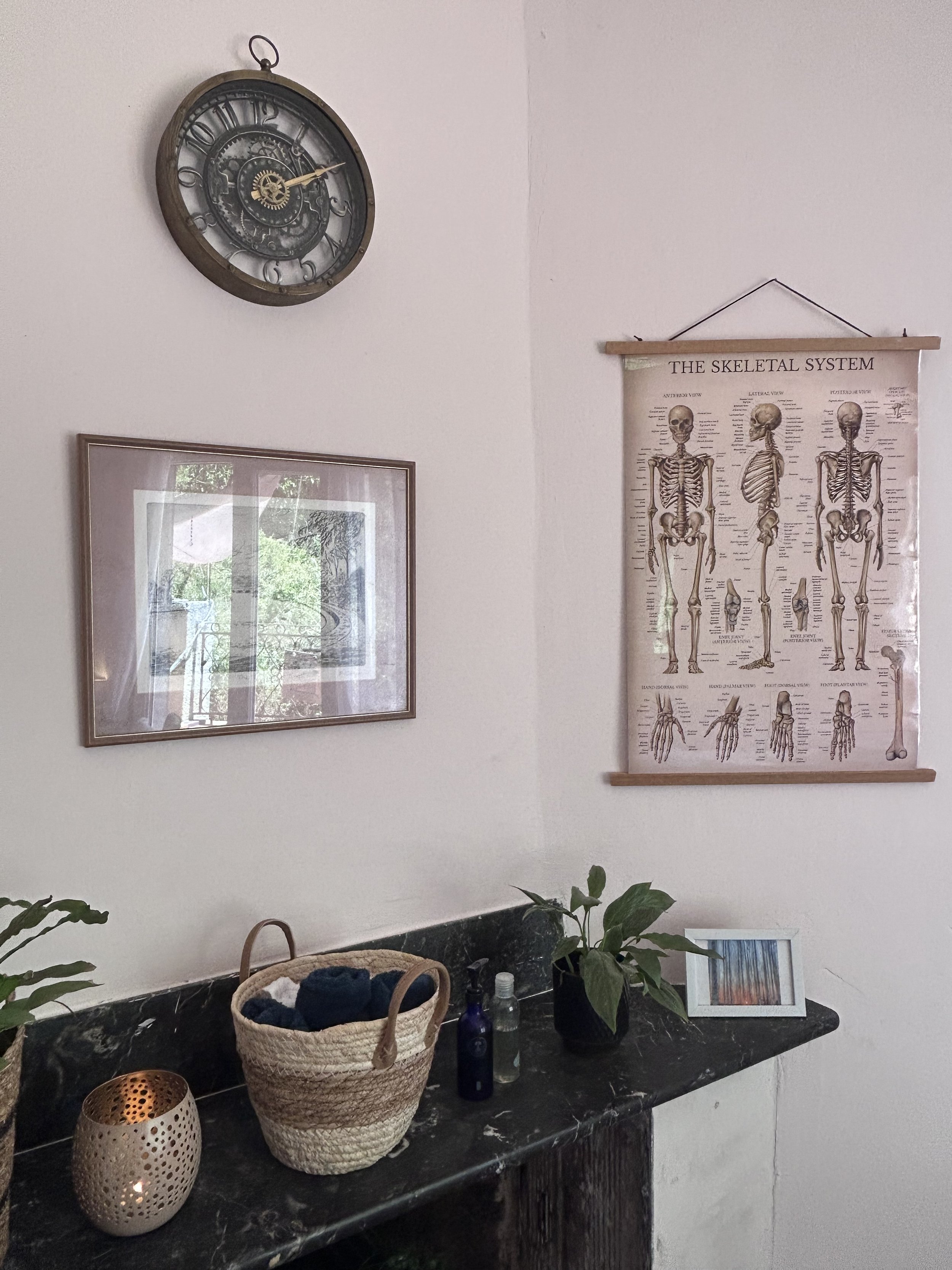 Décoration d'intérieur avec une horloge métallique vintage, un poster sur le système squelettique humain, un tableau encadré, un panier avec des tissus, des bouteilles en verre, une plante verte sur une surface en marbre noir.