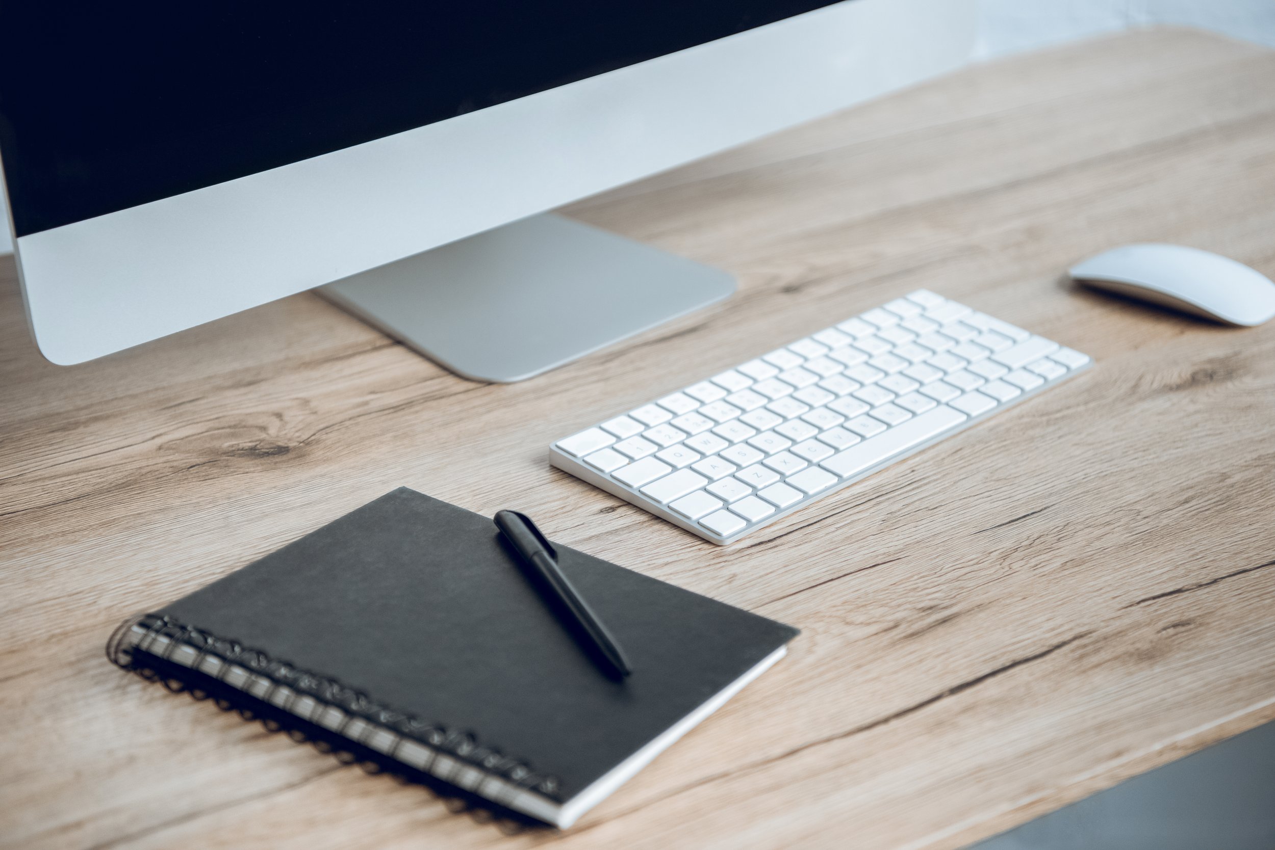 Bureau en bois avec un ordinateur, un clavier, une souris, un cahier noir, et un stylo noir.