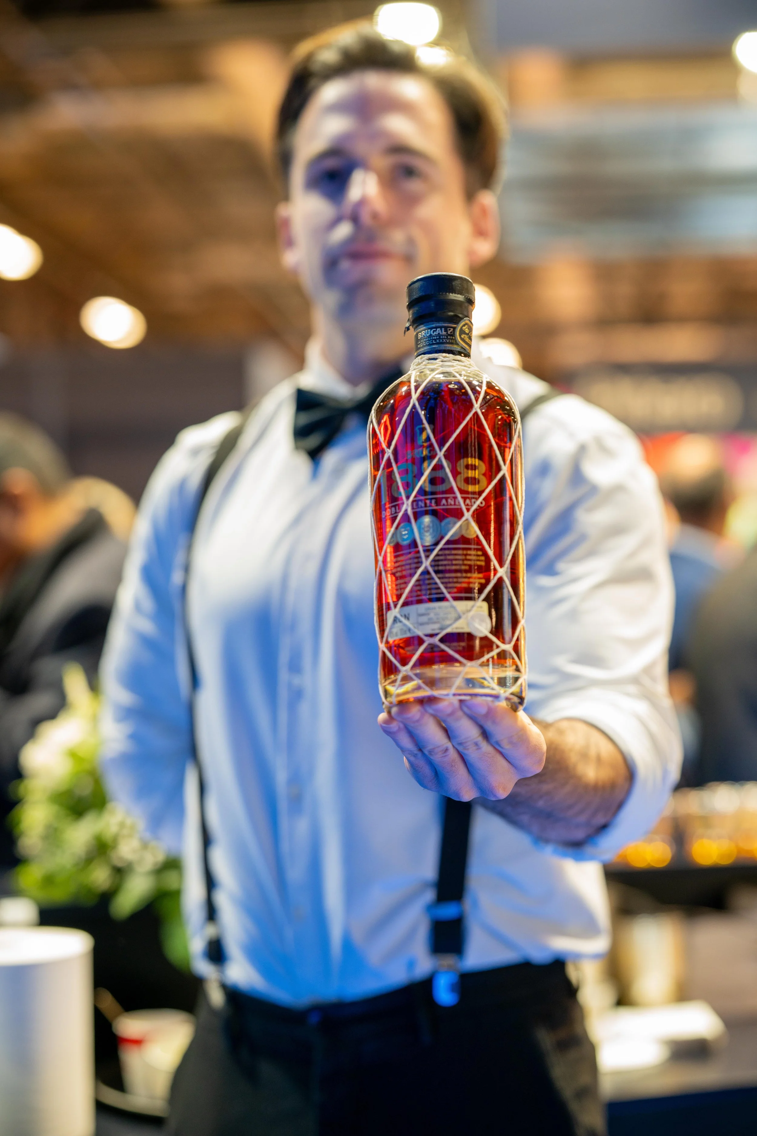  un homme en costume de serveur, avec une chemise blanche et un nœud papillon noir, tenant une bouteille de whisky devant lui dans un bar ou un restaurant animé.