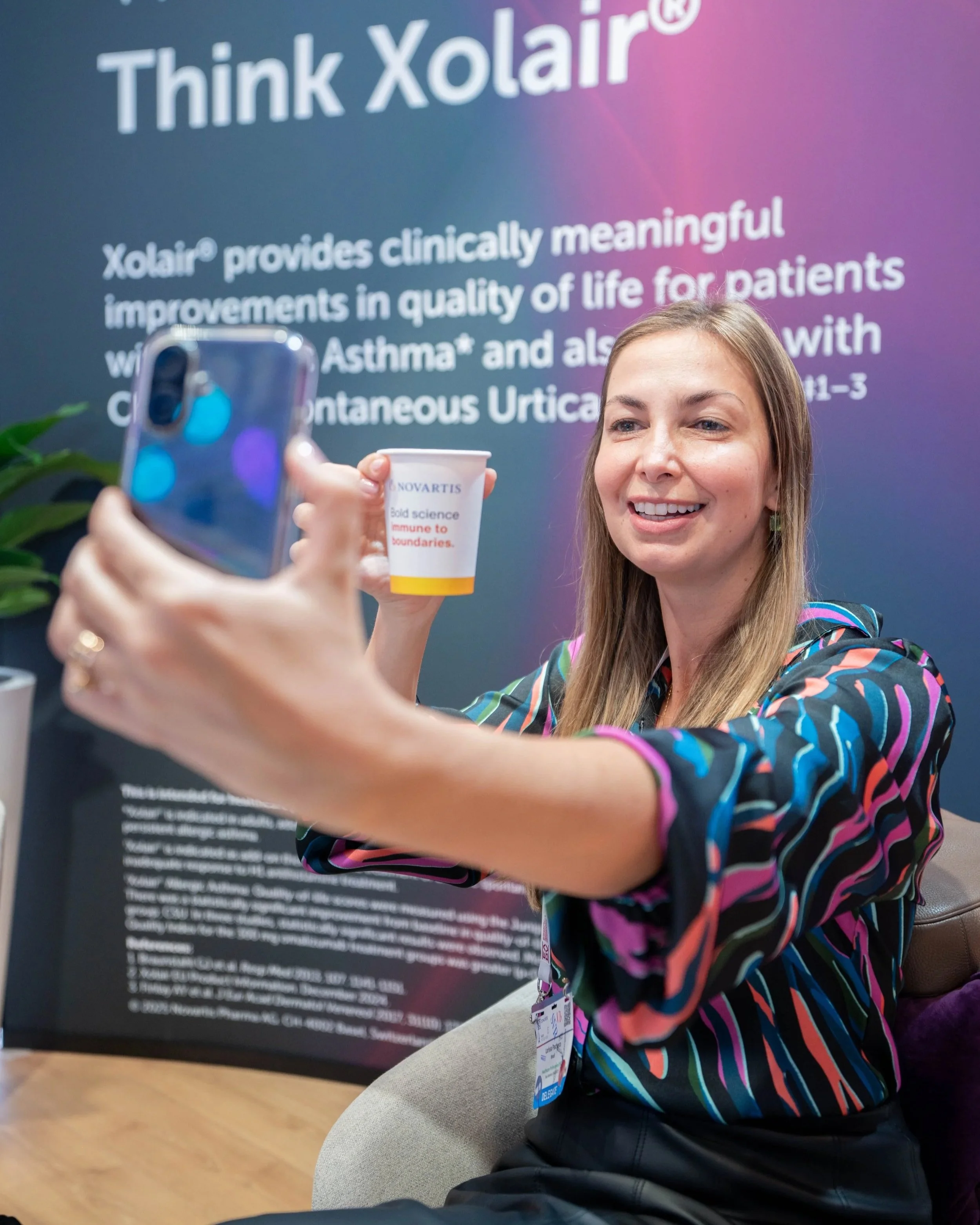 Une femme souriante photographie avec son téléphone une tasse blanche portant le logo de Novartis, dans un stand avec une affiche promotionnelle pour Xolair, un médicament destiné à améliorer la qualité de vie des patients souffrant d'asthme et d'urt