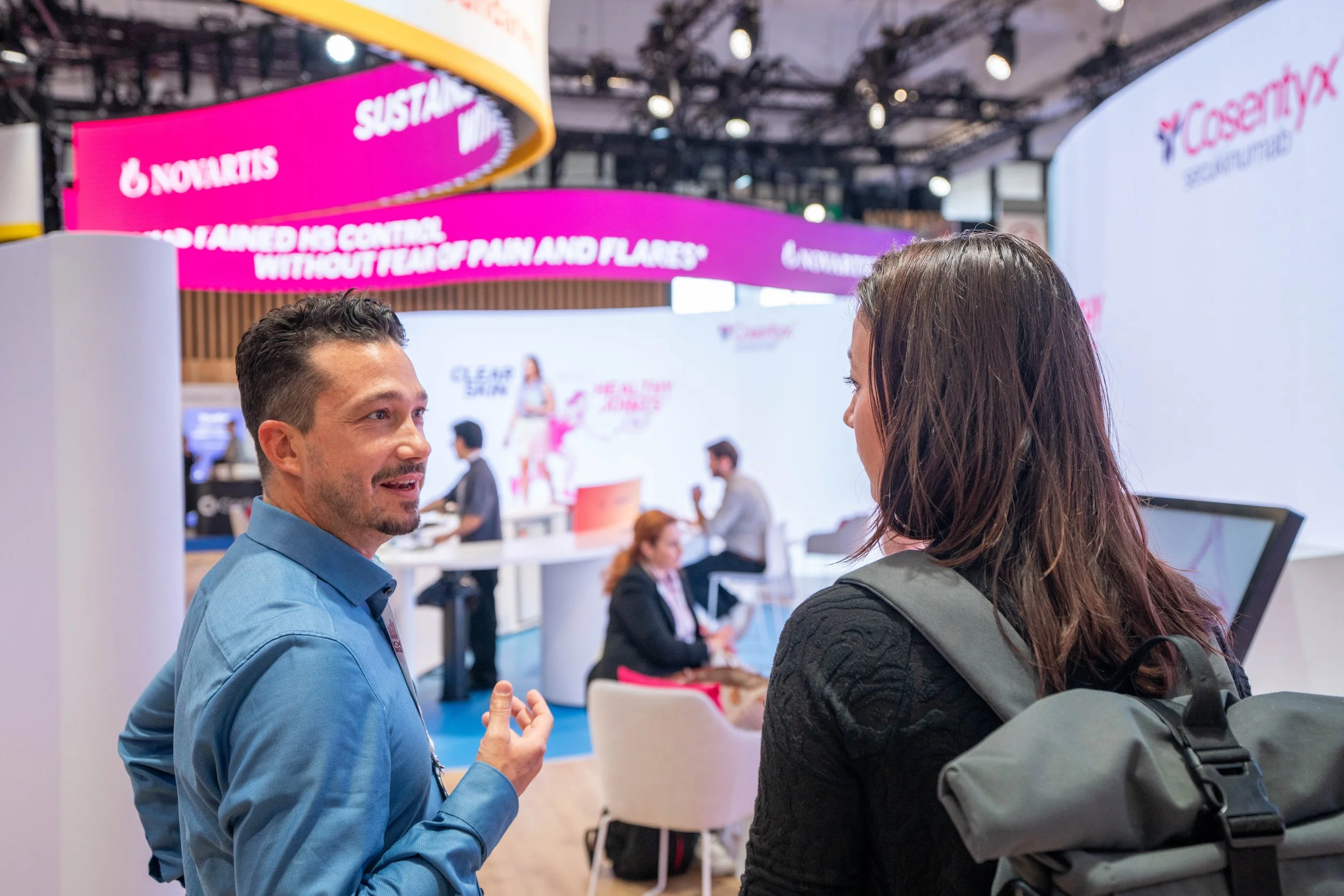 Deux personnes parlent dans un salon professionnel avec un grand écran derrière elles affichant des publicités pour un produit pharmaceutique.