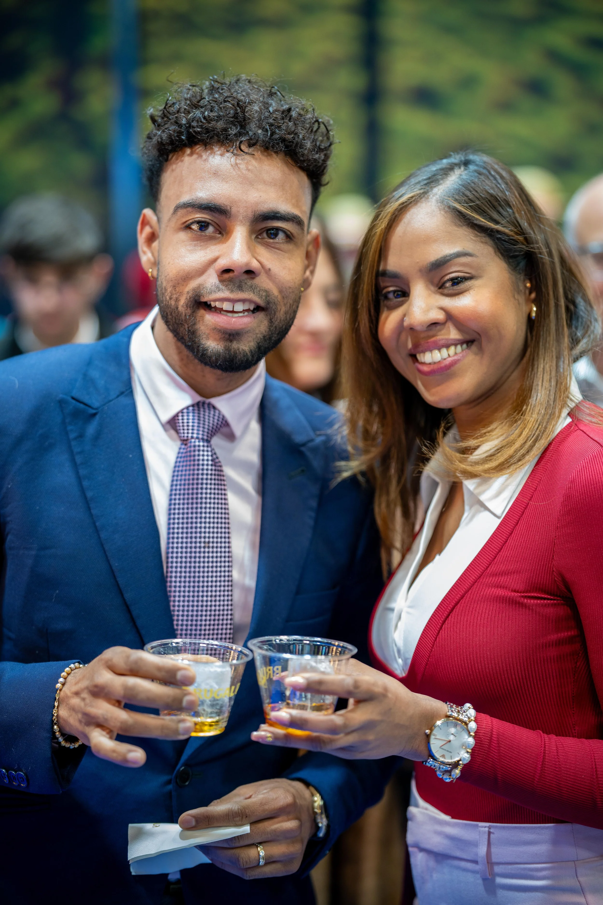 Deux personnes souriantes tenant des verres de verre à une fête ou un événement social, avec un arrière-plan flou de personnes et de nature.