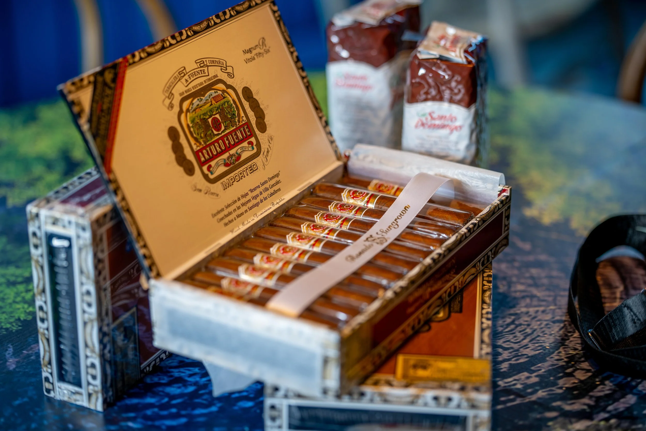 Une boîte ouverte de cigars Arturo Fuente contenant plusieurs cigars emballés, sur une table avec des paquets de sucre en arrière-plan et un ruban blanc à l'intérieur de la boîte.