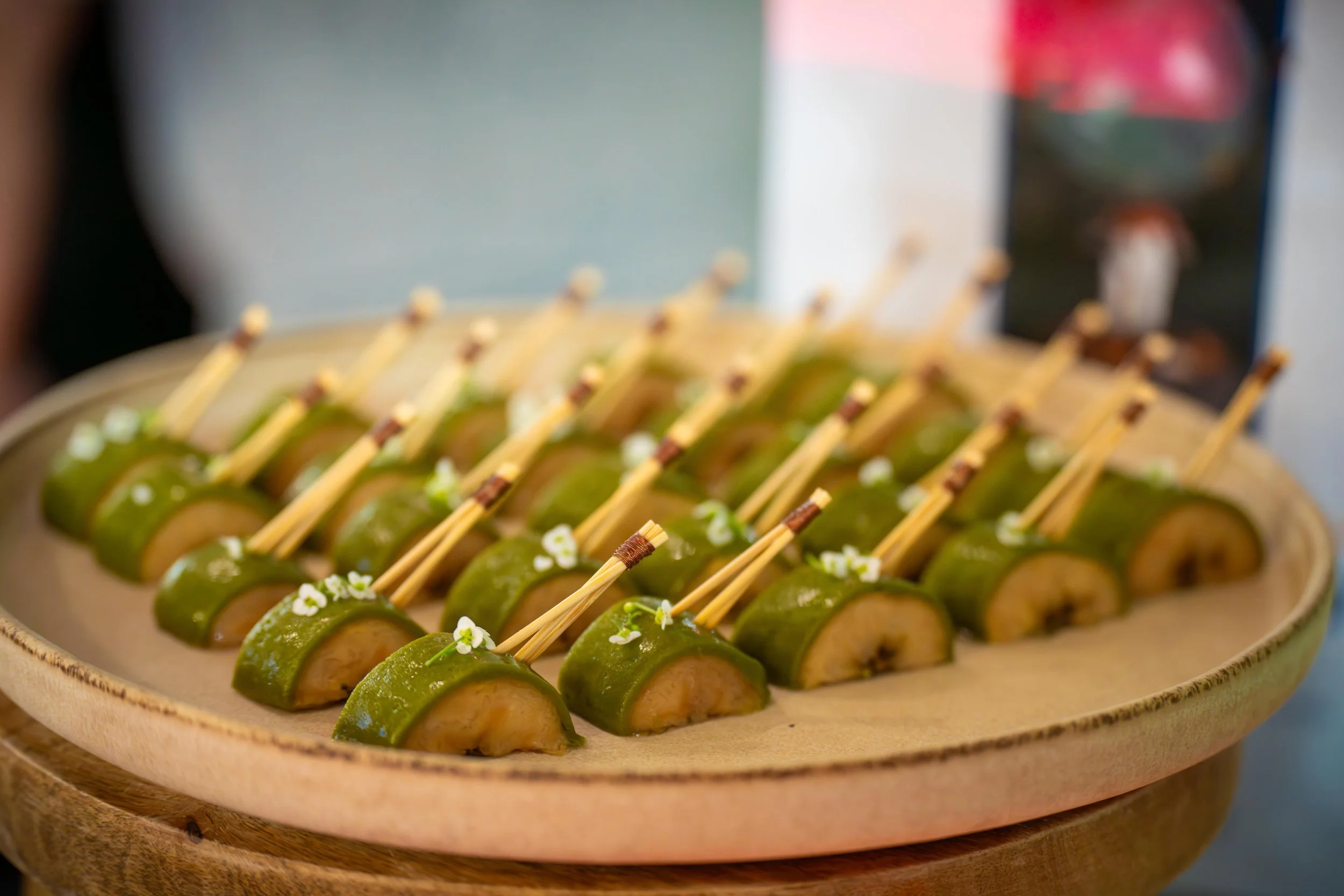 Bouchées de fruit vert avec un morceau de fruit à l'intérieur, piquées sur des petits brochettes, sur un plateau en bois, décorées de petites fleurs blanches.