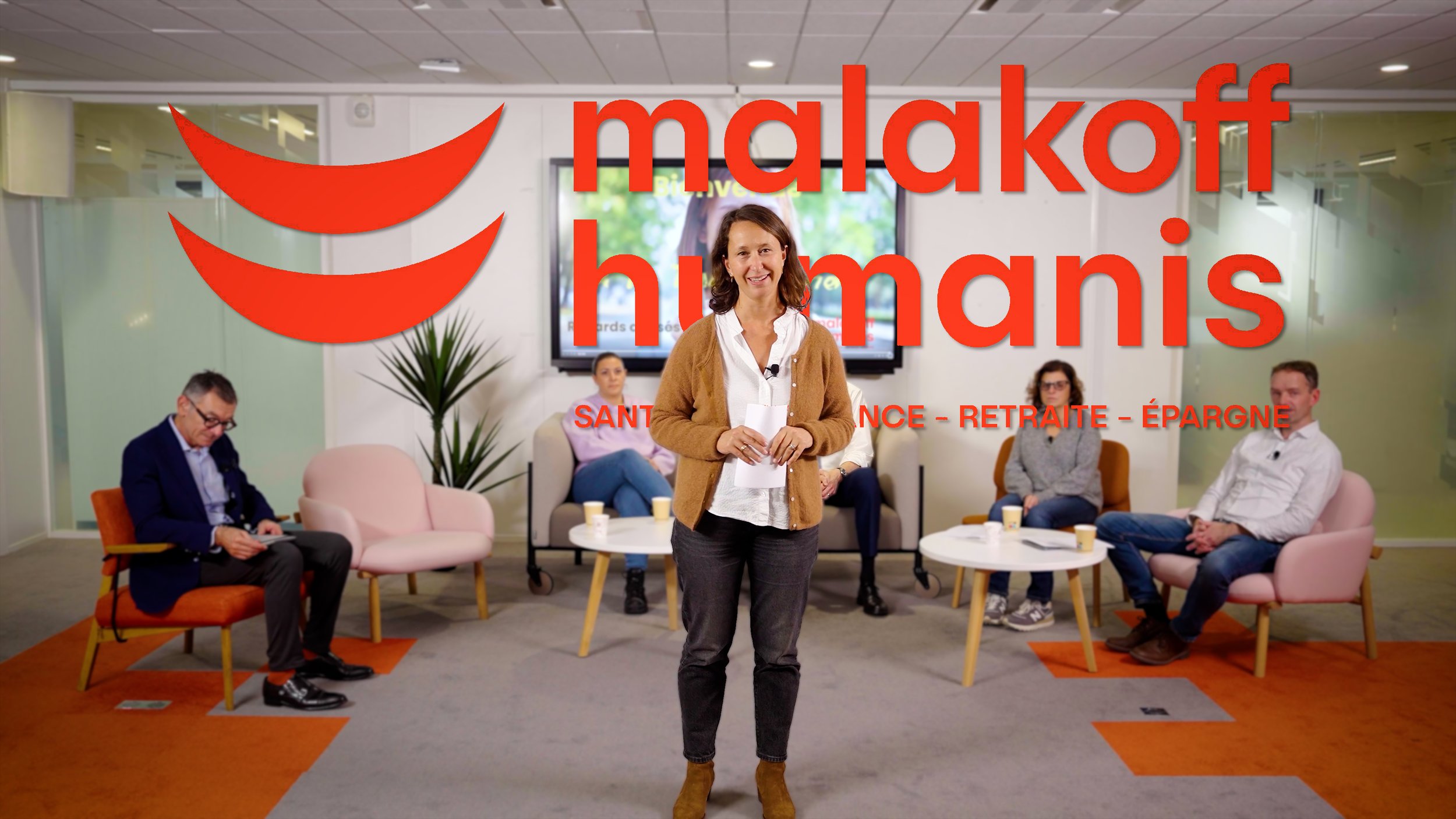 Une femme debout en studio, entourée de quatre personnes assises sur des fauteuils, avec un écran derrière affichant du texte. Sur l'image, il y a aussi un logo rouge en forme de deux croissants et le texte en français "malakoff humanis" avec d'autres mots comme "santé", "retraite" et "épargne".