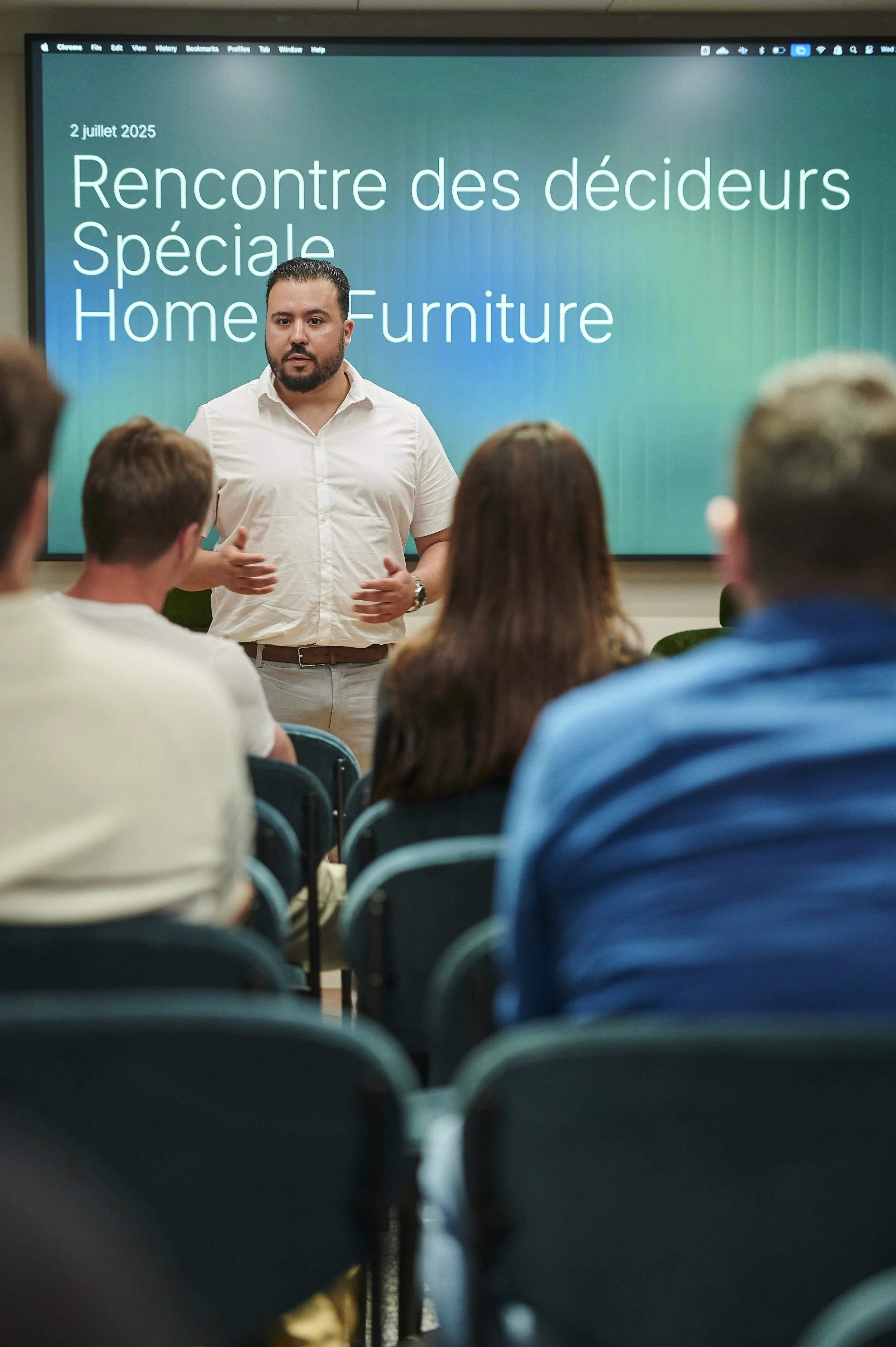 Un homme en chemise blanche donne une présentation à un groupe de personnes dans une salle, avec un écran affichant la date '2 juillet 2025' et le texte 'Rencontre des décideurs Spéciale Home Furniture'.