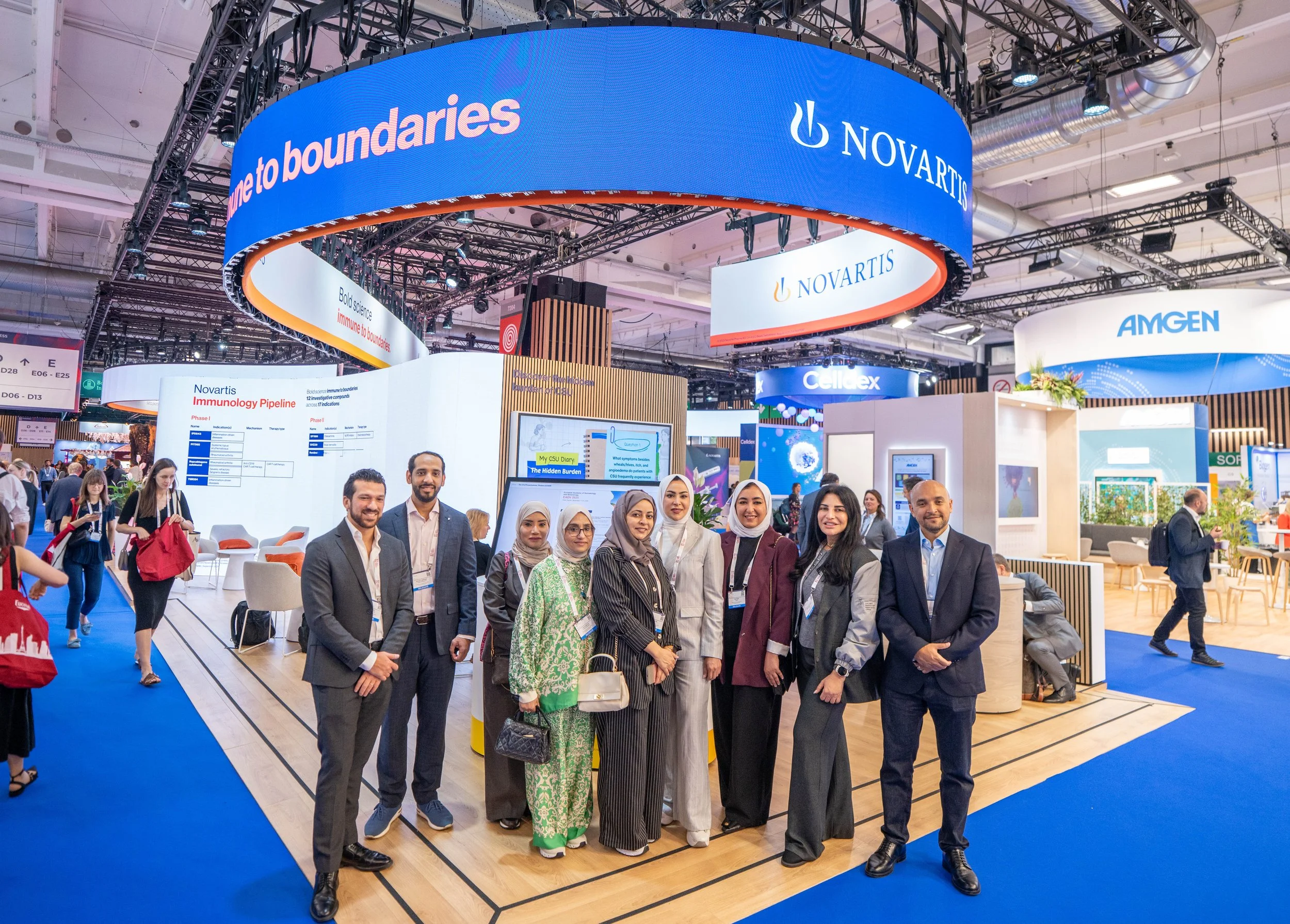 Groupe de personnes posant dans un stand d'exposition avec des affiches et des écrans, sous une grande bannière bleue avec le logo NOVARTIS