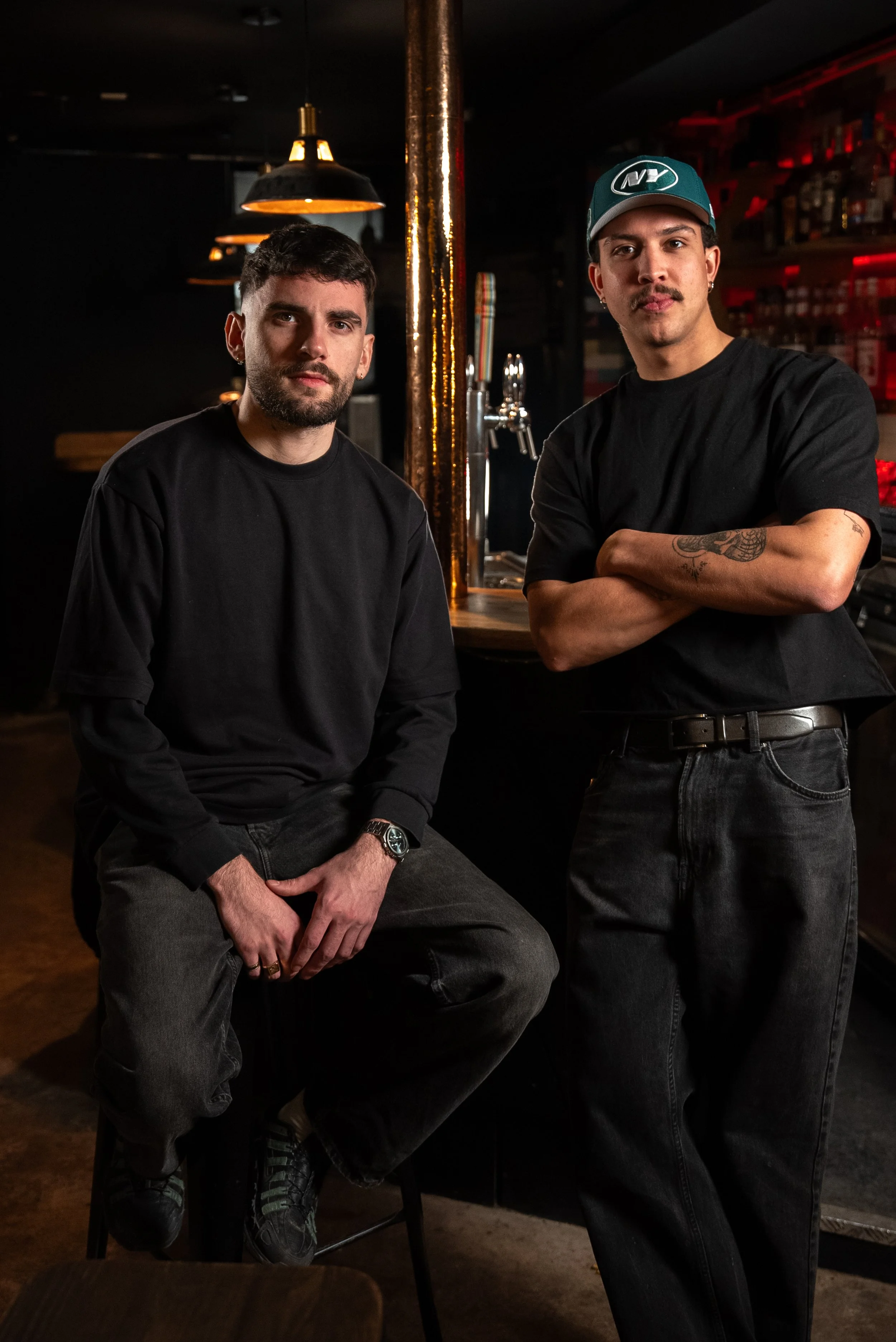 Deux jeunes hommes dans un bar ou un café, l'un assis sur une chaise avec les jambes croisées et l'autre debout, les bras croisés. L'ambiance est sombre avec un éclairage chaud et des étagères derrière eux.