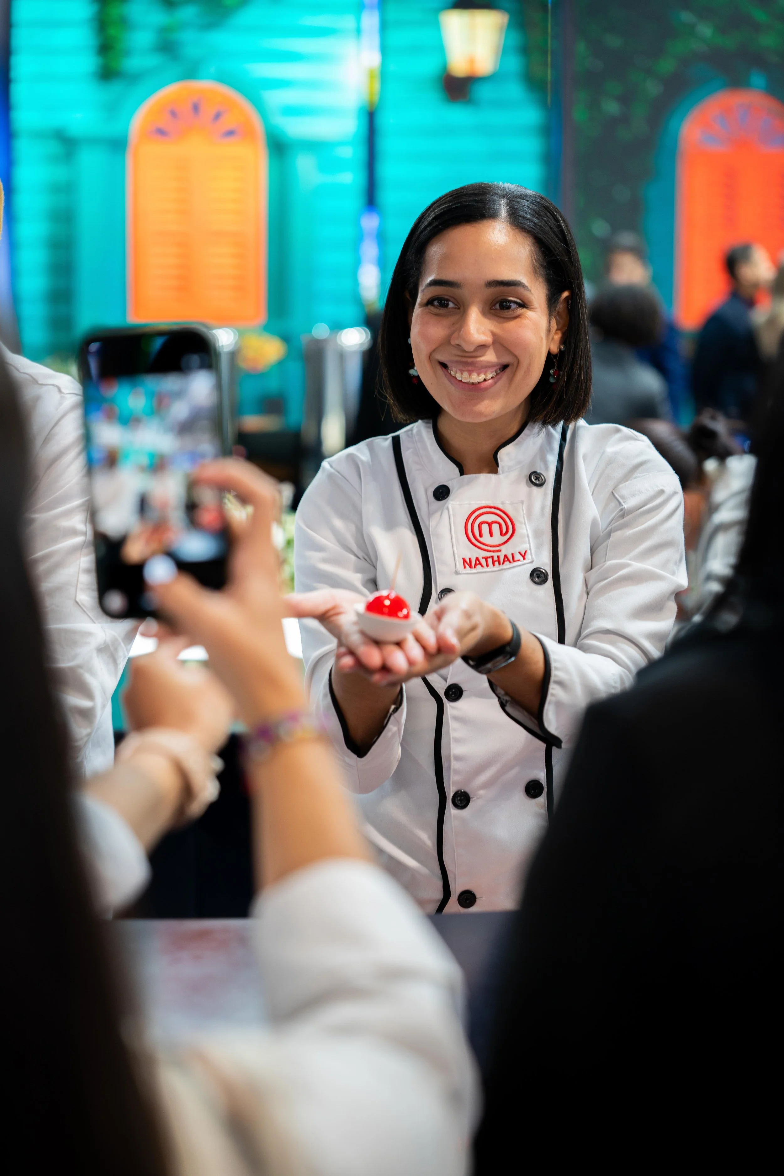 Une femme chef en uniforme de cuisine, avec le nom Nathaly, tenant un dessert rouge avec une cerise dessus, lors de la prise de photo par une personne dans un restaurant ou une cuisine. En arrière-plan, un décor coloré avec des fenêtres orange et des