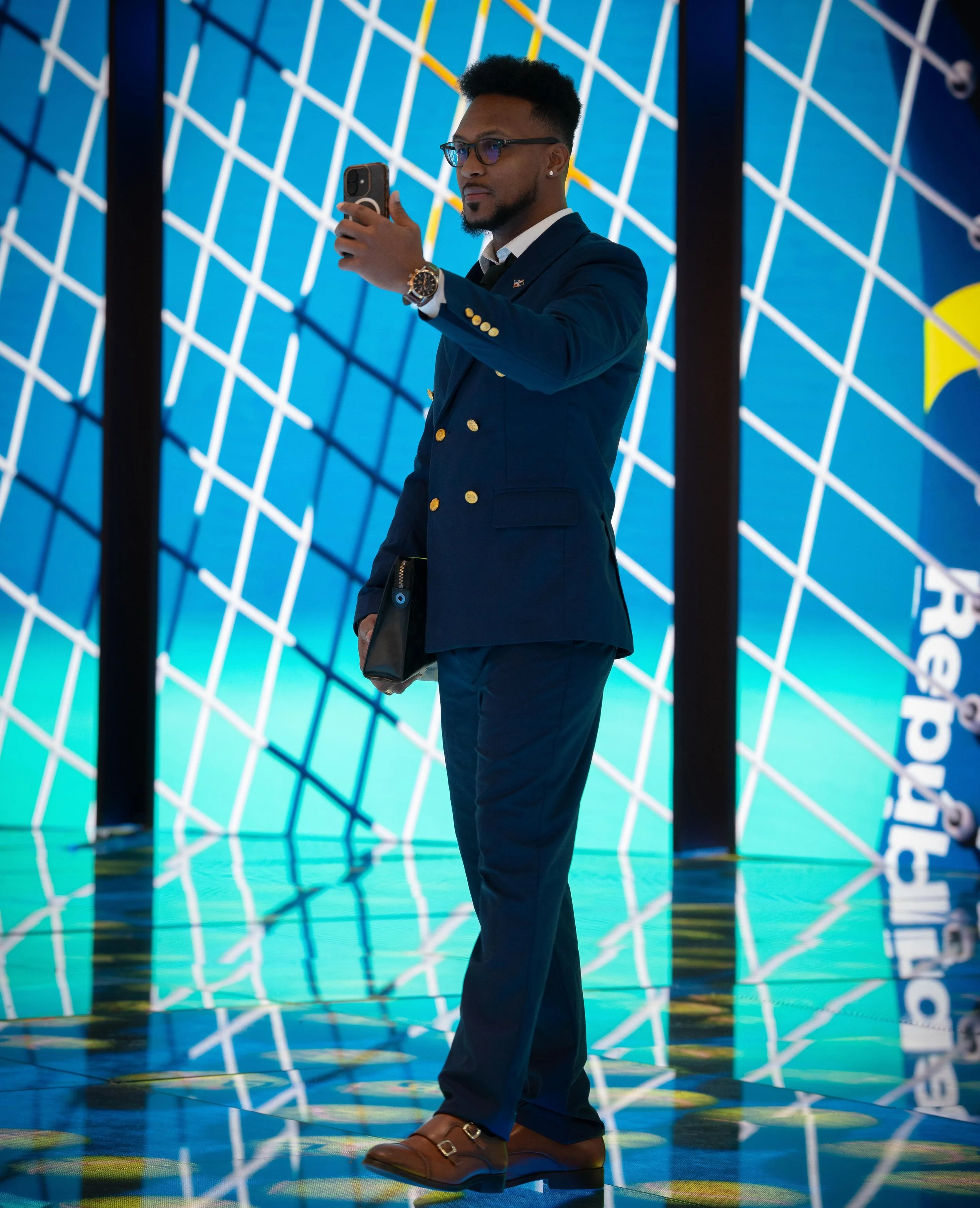 Un homme d'affaires en costume bleu, portant des lunettes, coiffé de cheveux courts et coiffés, doit tenir un téléphone portable en train de prendre une photo ou de faire une vidéo. Il porte une montre et une porte-documents. L'arrière-plan est un mu