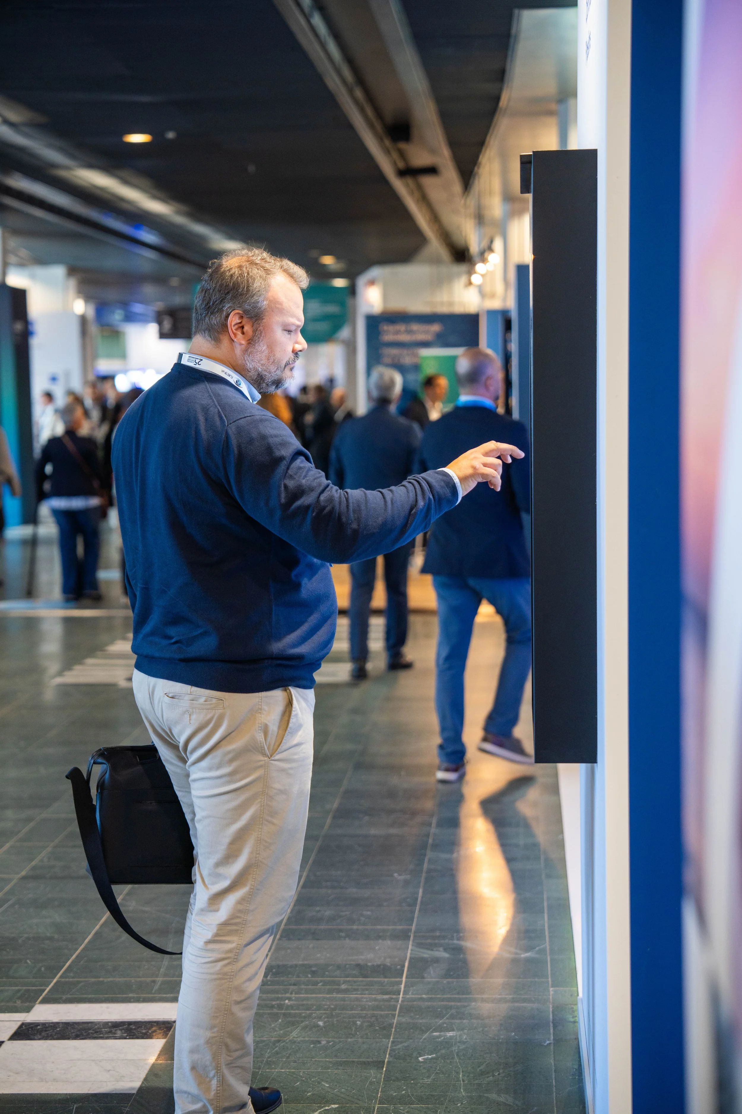 Un homme d'âge moyen avec une barbe, portant un sweat bleu et un pantalon beige, se tient debout dans une salle de conférence ou un salon professionnel. Il interagit avec un écran tactile fixé au mur, dans une foule de personnes en arrière-plan qui d