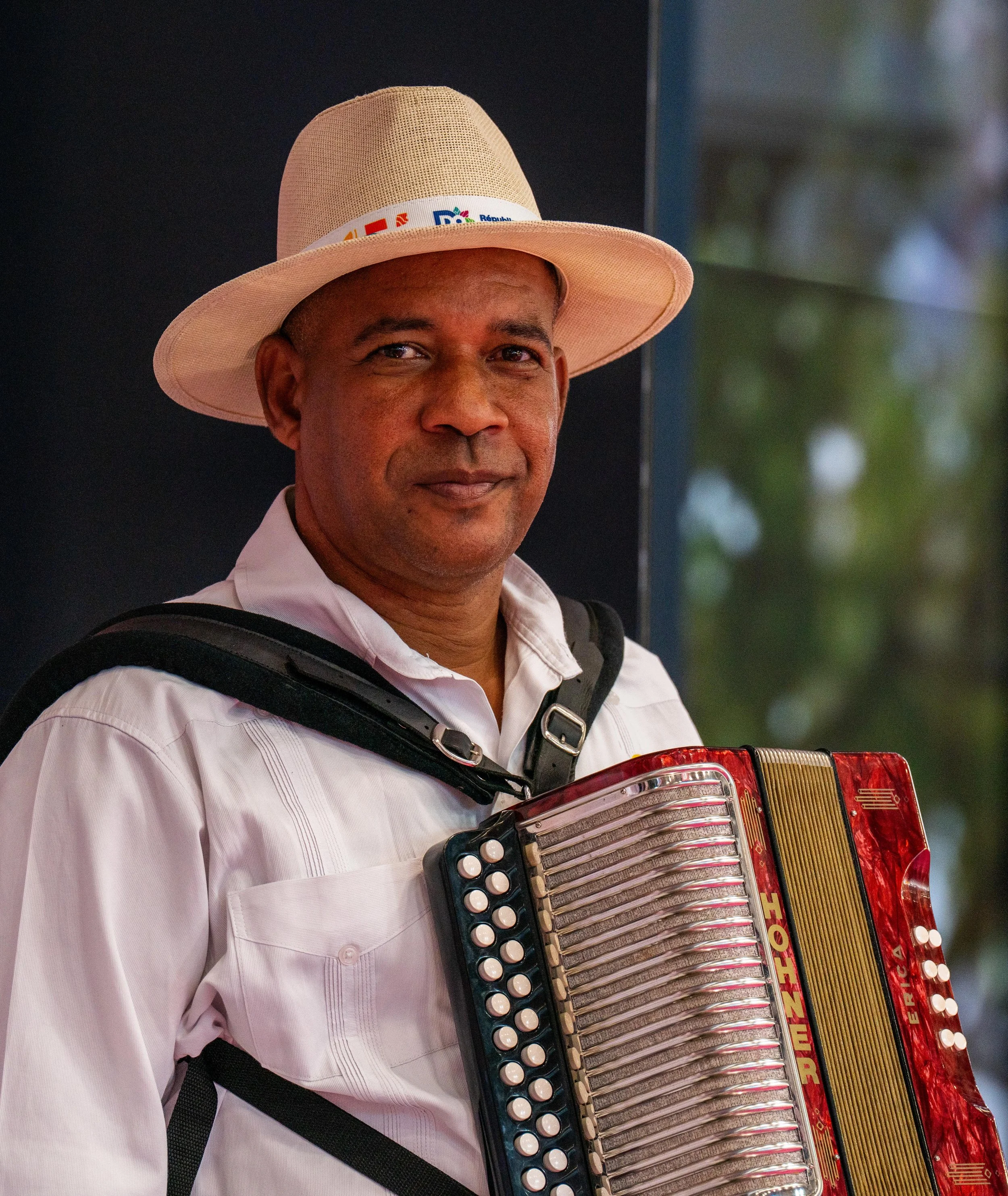 Homme de profil portant un chapeau blanc, une chemise blanche et jouant de l'accordéon, avec un fond flou et un mur bleu.