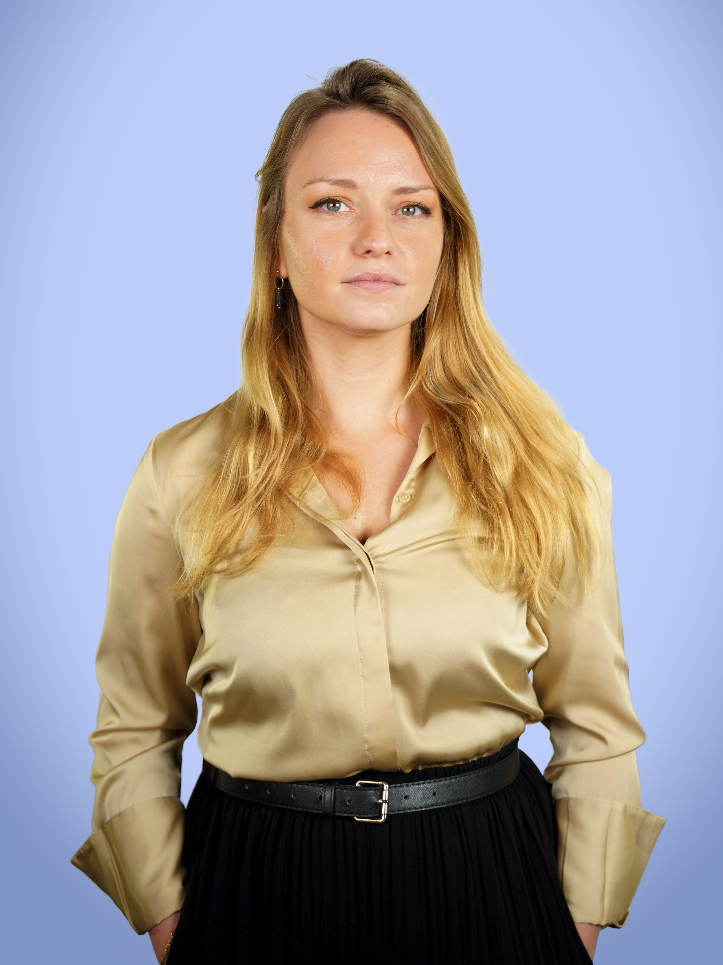 Jeune femme aux cheveux longs blond, portant une chemise beige en satin et une jupe noire, avec un fond bleu clair.