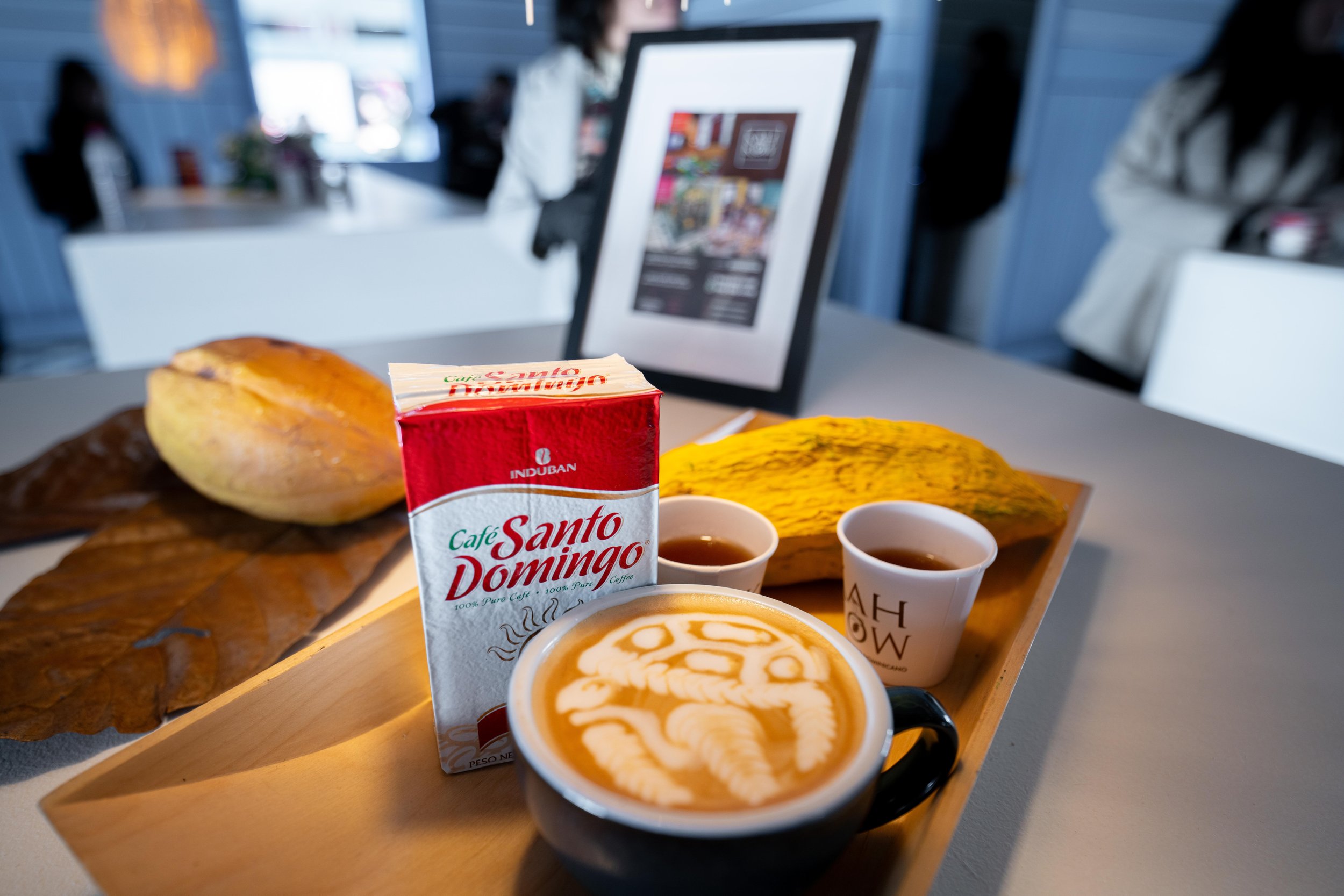 Un plateau avec une tasse de café latte avec de la crème artistique, deux petites tasses de café noir, une boîte de café Santo Domingo, un morceau de sucre coloré, une banane mûre, et une feuille sèche sur une table dans un café moderne.