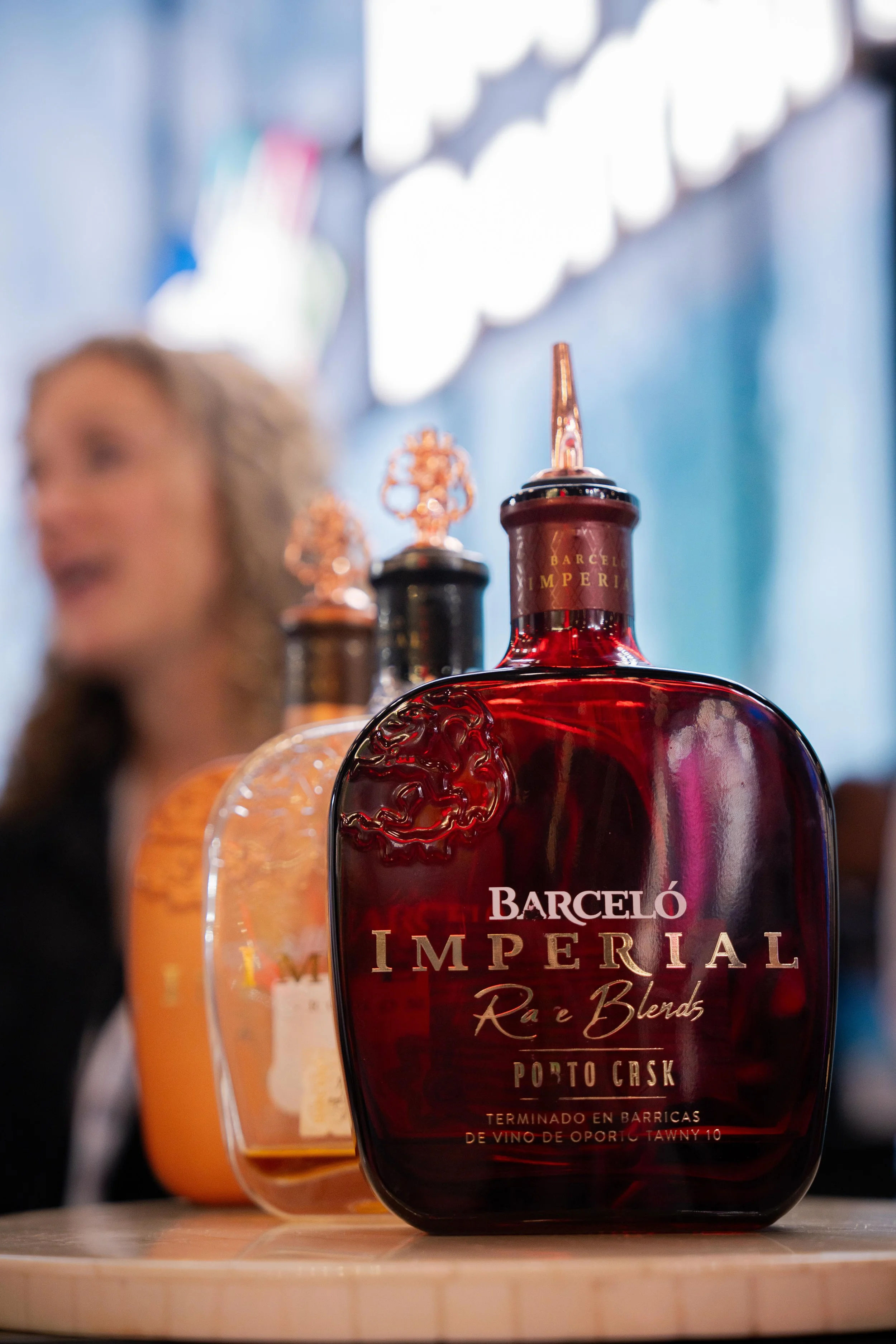 Une bouteille rouge de Porto Cask Barceló Imperial Rye & Blends en premier plan, derrière d'autres bouteilles et un flacon, avec une femme floue en arrière-plan dans un environnement intérieur.