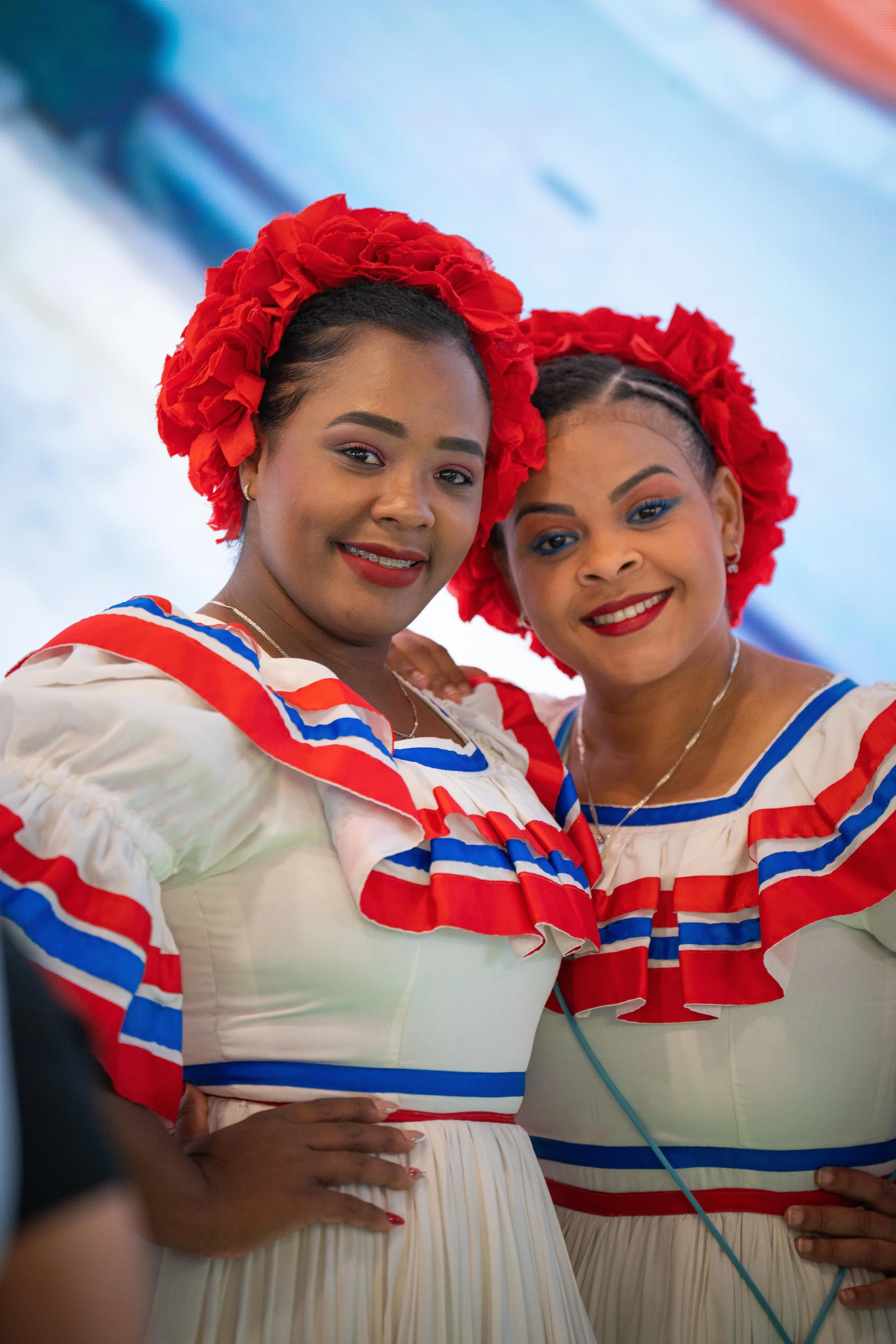 Deux femmes portent des tenues traditionnelles avec des ruffles rouges, bleus et blancs, et des coiffures ornées de fleurs rouges.