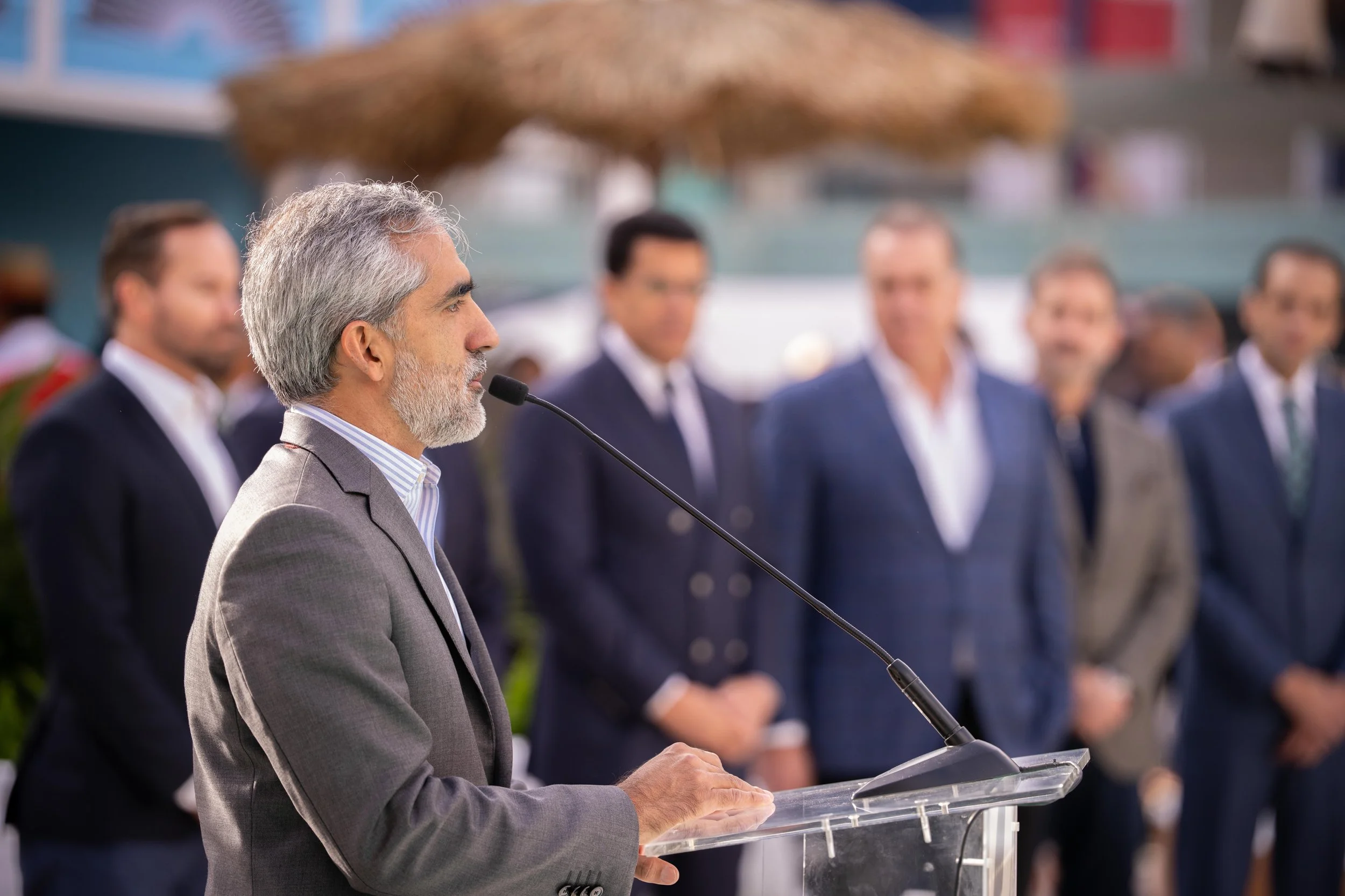 Un homme à cheveux gris et barbe parle lors d'un discours à un micro, entouré de plusieurs personnes en costume lors d'un événement en plein air.