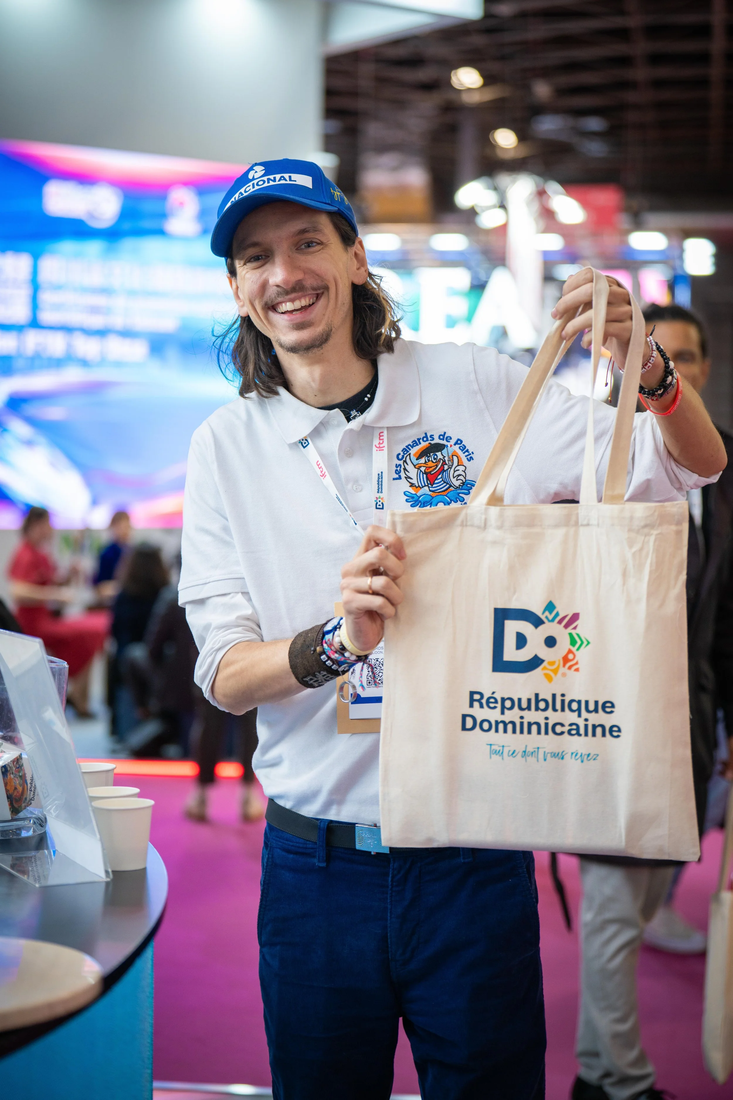 Un homme souriant tenant un sac en tissu avec le logo de la République Dominicaine, portant un polo blanc avec un badge, dans un environnement d'événement ou de conférence.