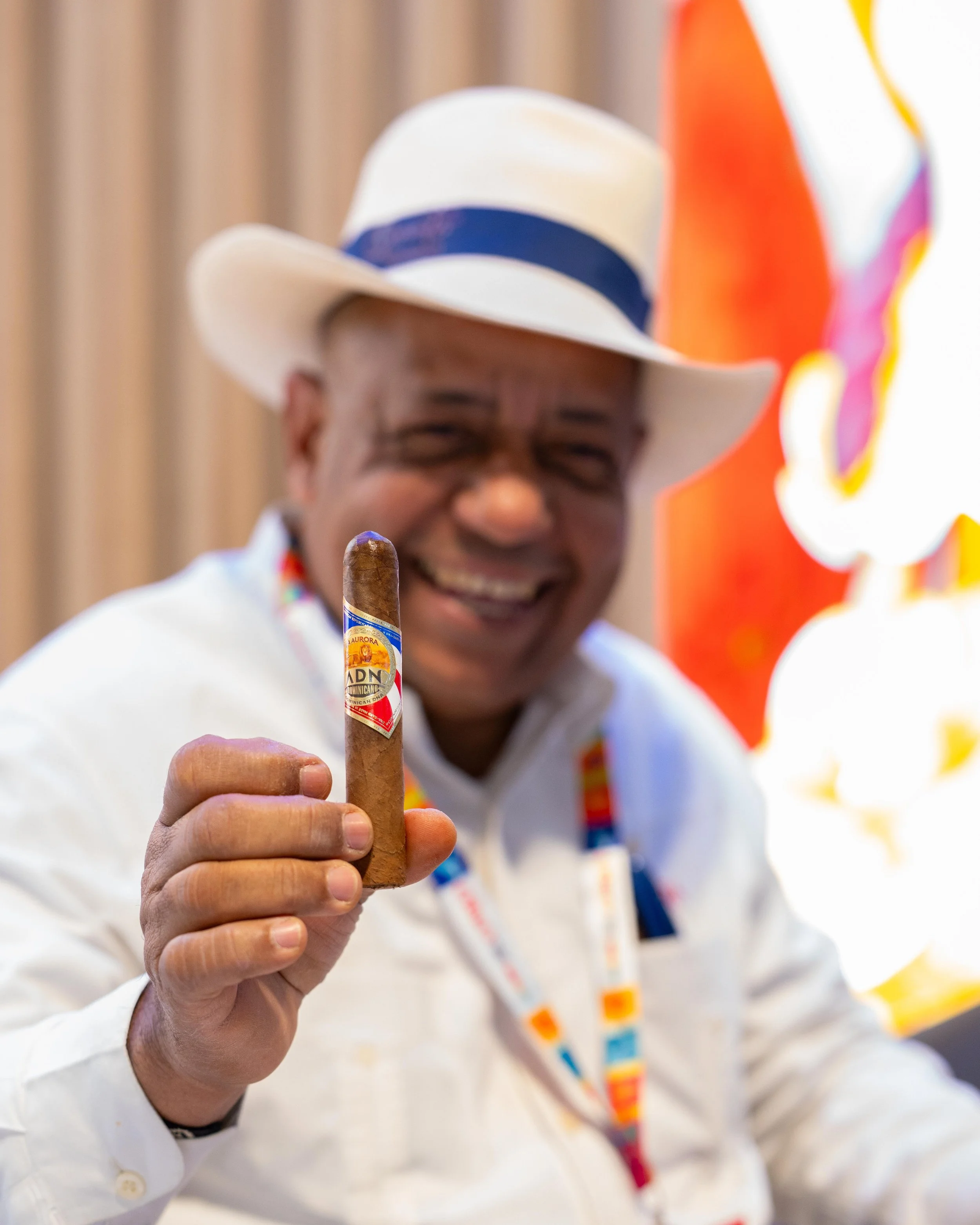 Homme souriant portant un chapeau blanc et une chemise blanche, tenant un cigare avec une étiquette colorée, dans un environnement coloré et vibrant.