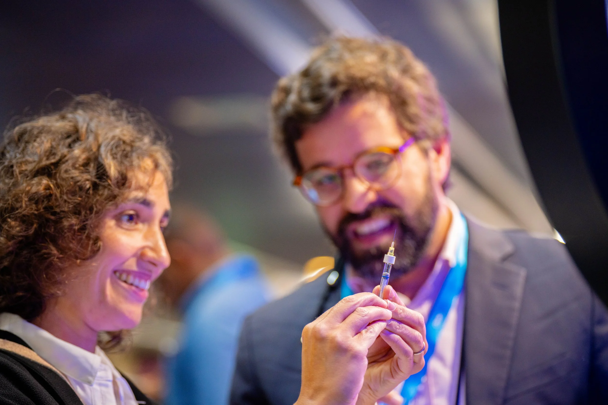 Deux personnes, un homme et une femme, discutent lors d'une conférence. La femme sourit et regarde l'homme, qui tient une seringue et semble expliquer quelque chose. L'ambiance est professionnelle et conviviale.