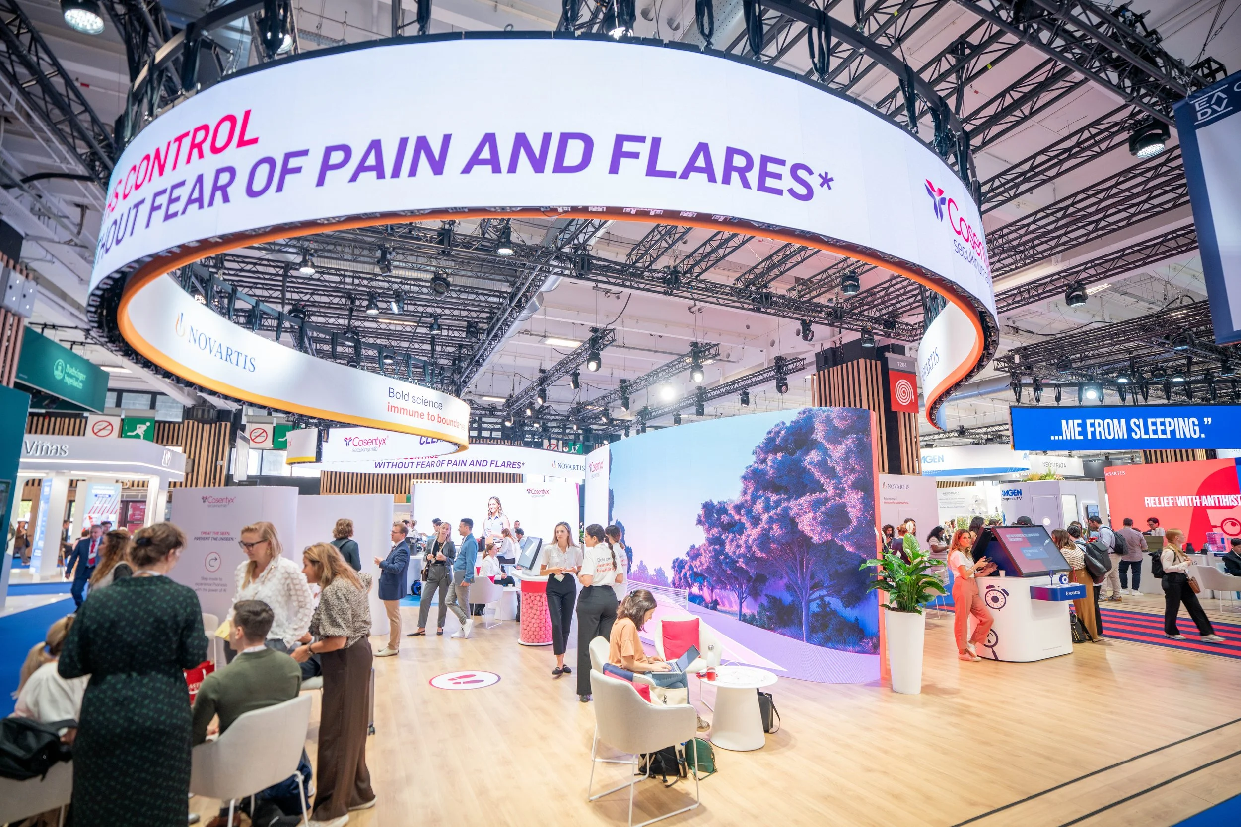 Une scène d'exposition avec plusieurs stands, une grande bannière circulaire affichant le texte "CONTROL YOUR FEAR OF PAIN AND FLARES", et des visiteurs discutant et explorant les stands.