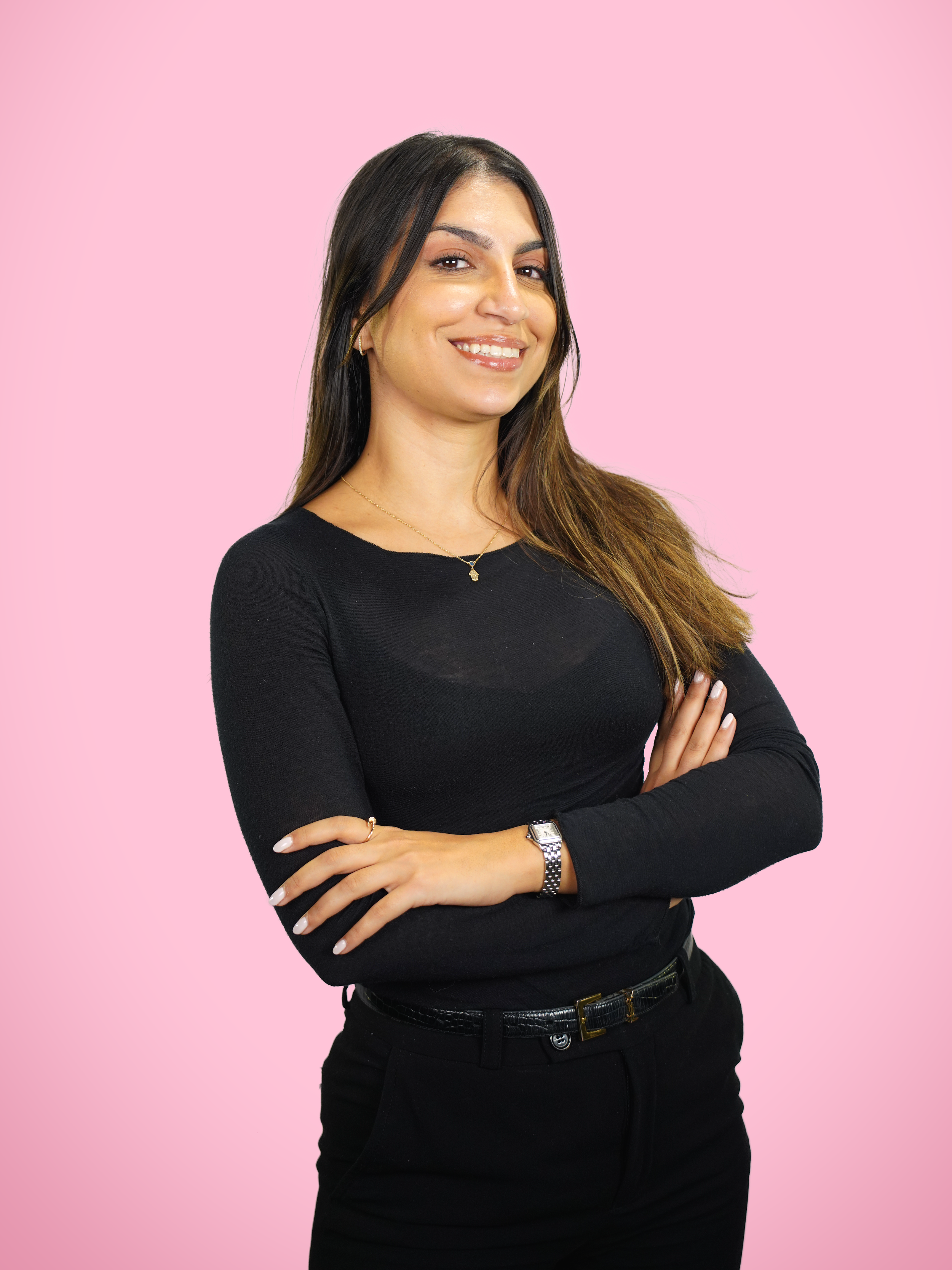 Une femme souriante, portant un haut noir, des bijoux et une montre, posant avec les bras croisés contre un fond rose.