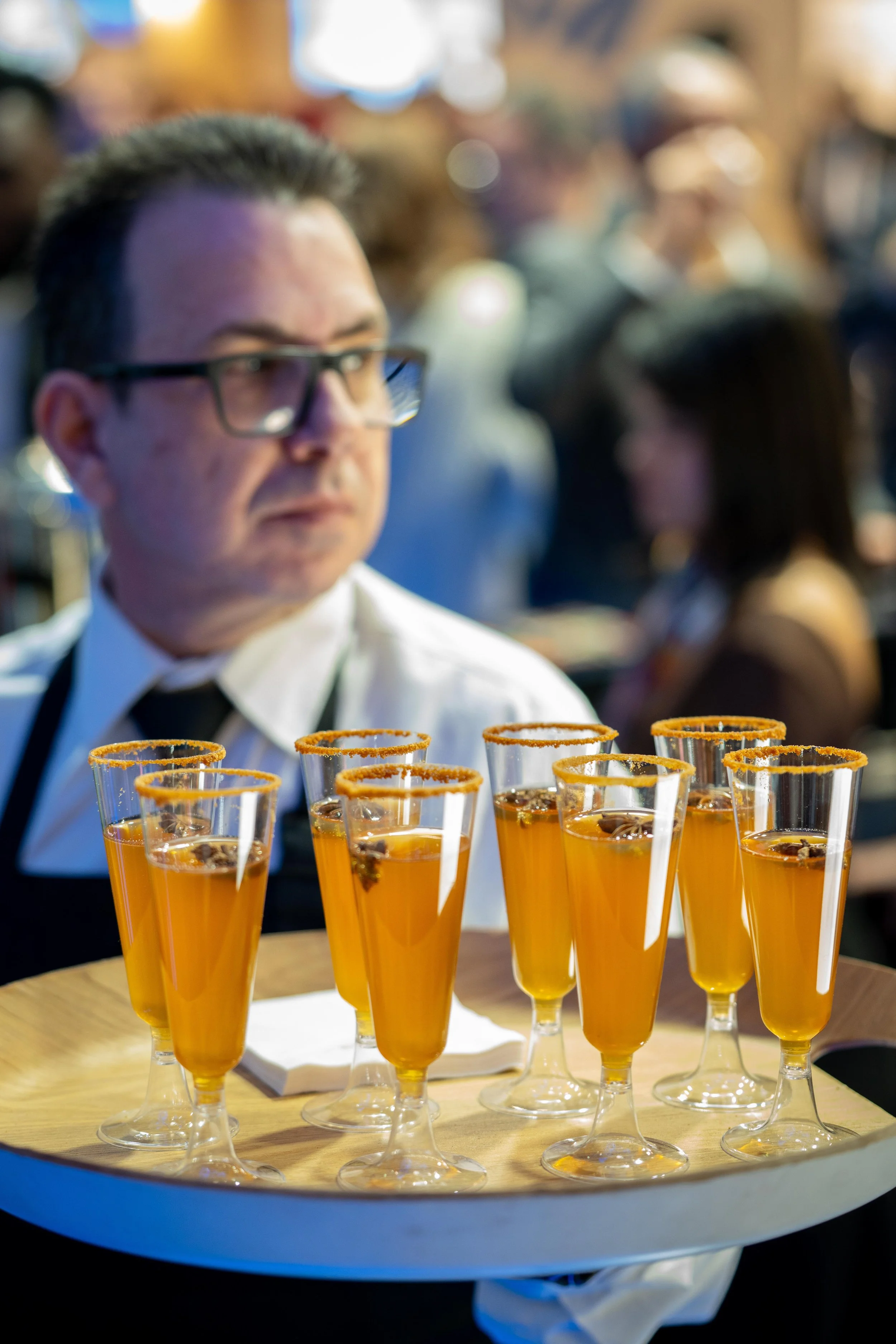 Homme portant des lunettes et une chemise blanche, servant des cocktails en flûtes avec un rebord épicé, dans un environnement animé.