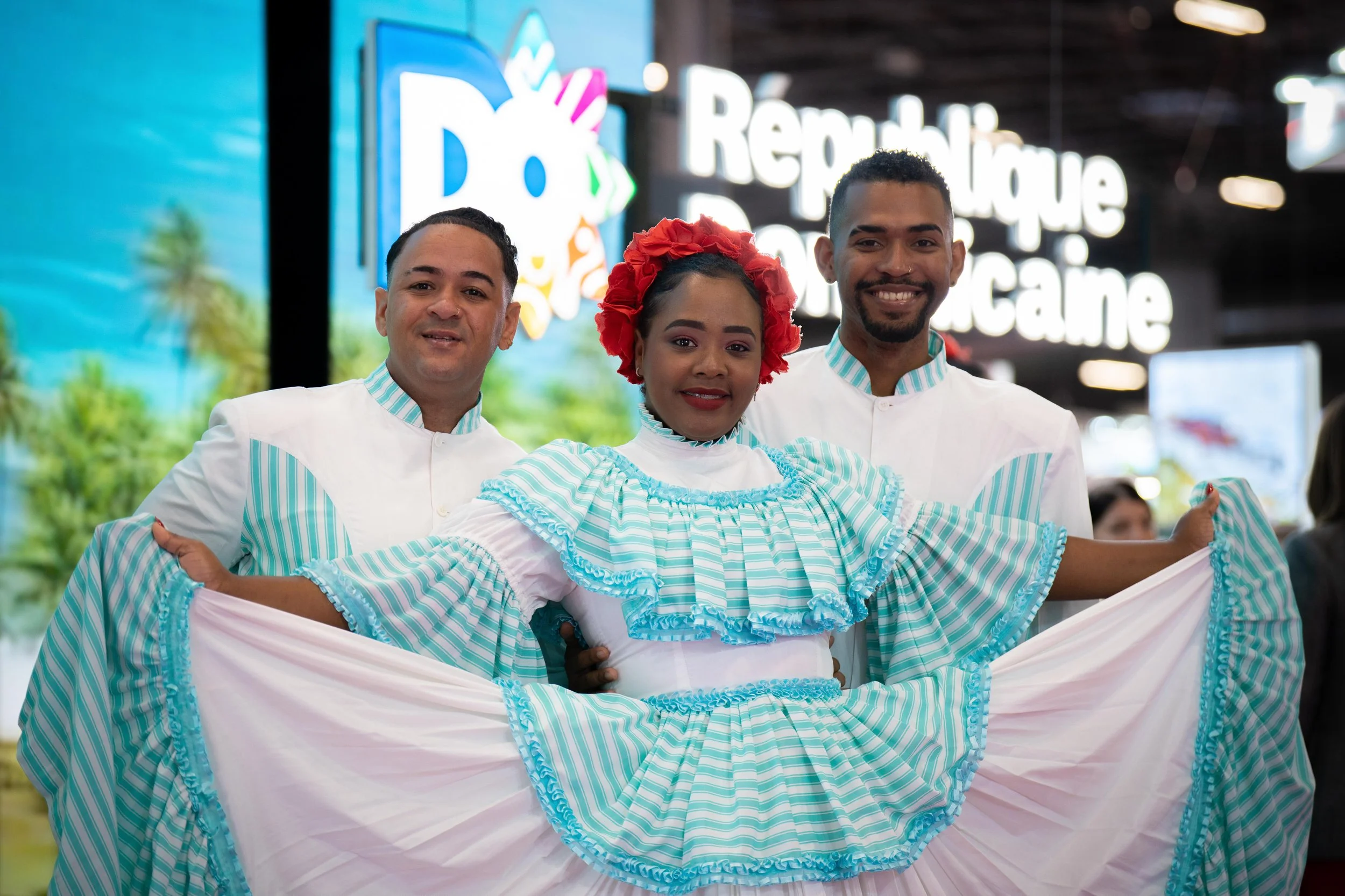 Trois personnes vêtues de costumes traditionnels colorés, deux hommes en chemises blanches et une femme avec une robe à rayures bleues et blanches, souriantes, posant dans un centre commercial.