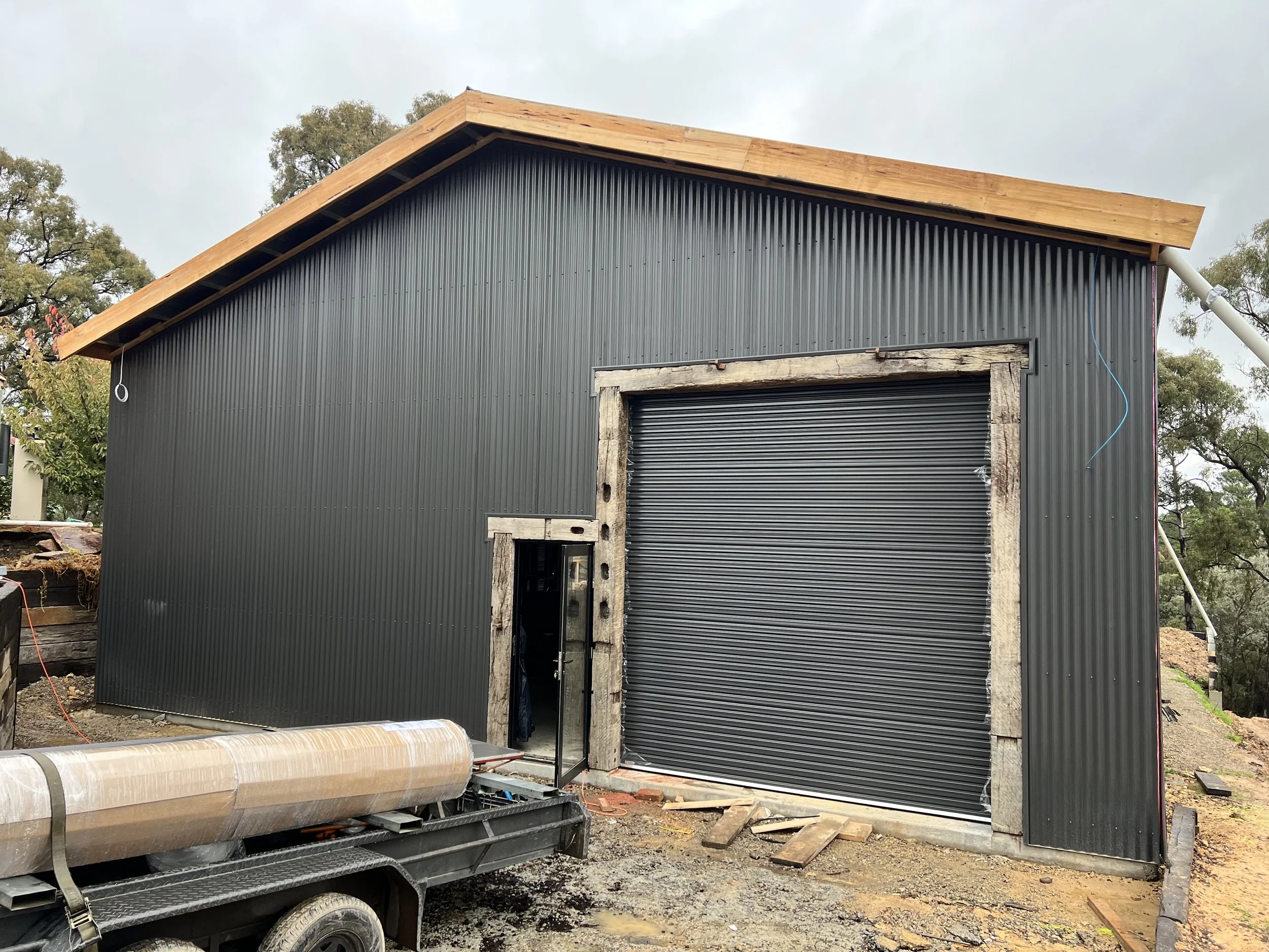 A building under construction with black corrugated metal siding, a large roll-up garage door, and a partially finished wooden roof framework. There is construction equipment in front and trees nearby.
