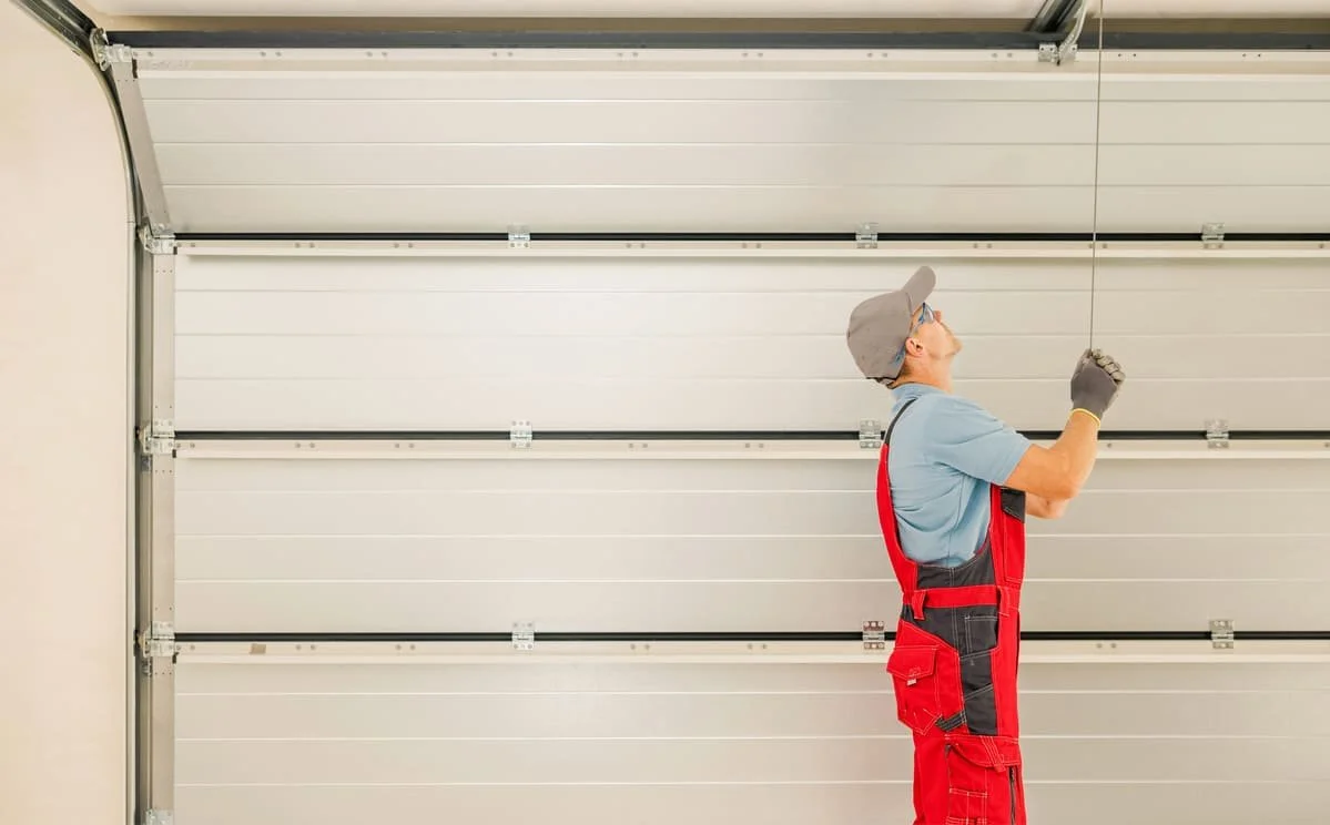 A person in red work overalls, a gray cap, and gloves standing on a ladder, reaching up to open a garage door.
