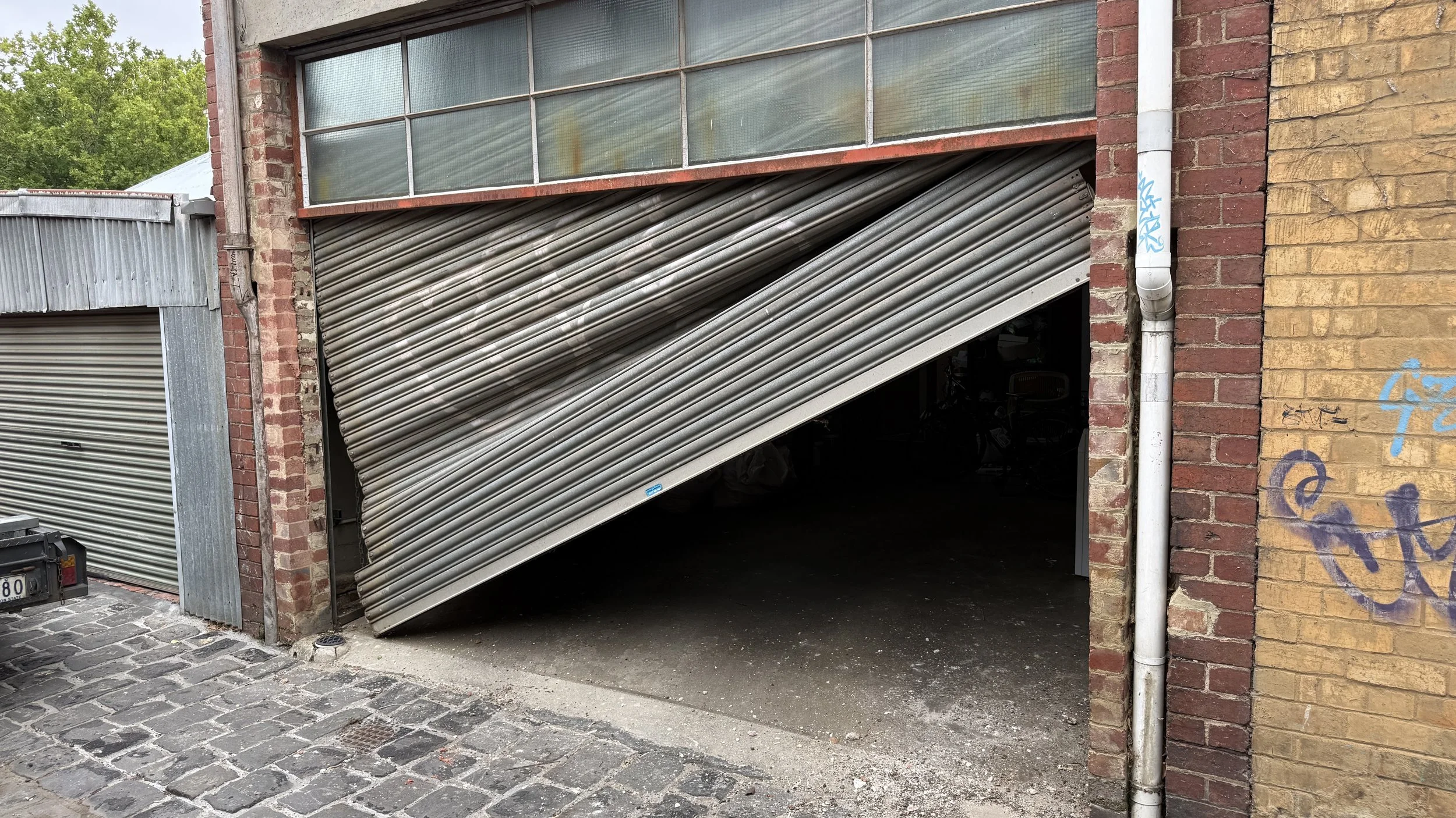 A partially open metal garage door on a brick building, with a smaller door to its left and graffiti on the yellow brick wall to the right.