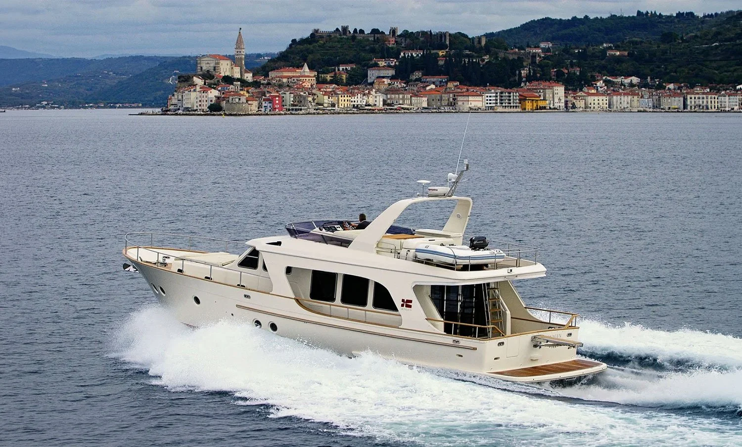 A white yacht moving through the water with a city and hillside town in the background.