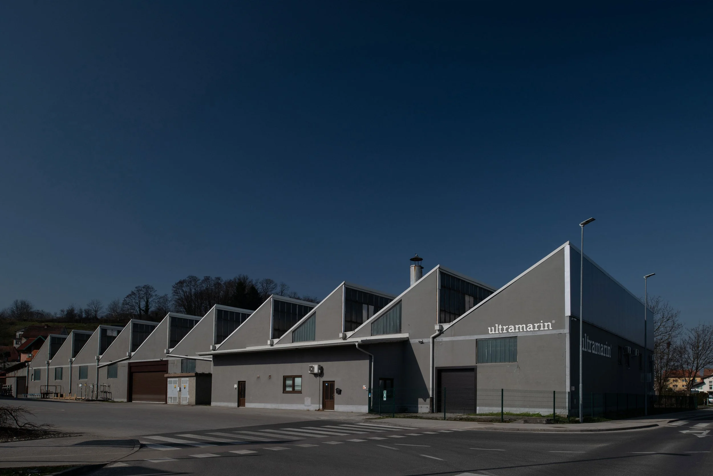 An industrial building with a gray exterior and multiple triangular rooftops, labeled 'ultramarin,' on a street corner during daytime with a clear blue sky.