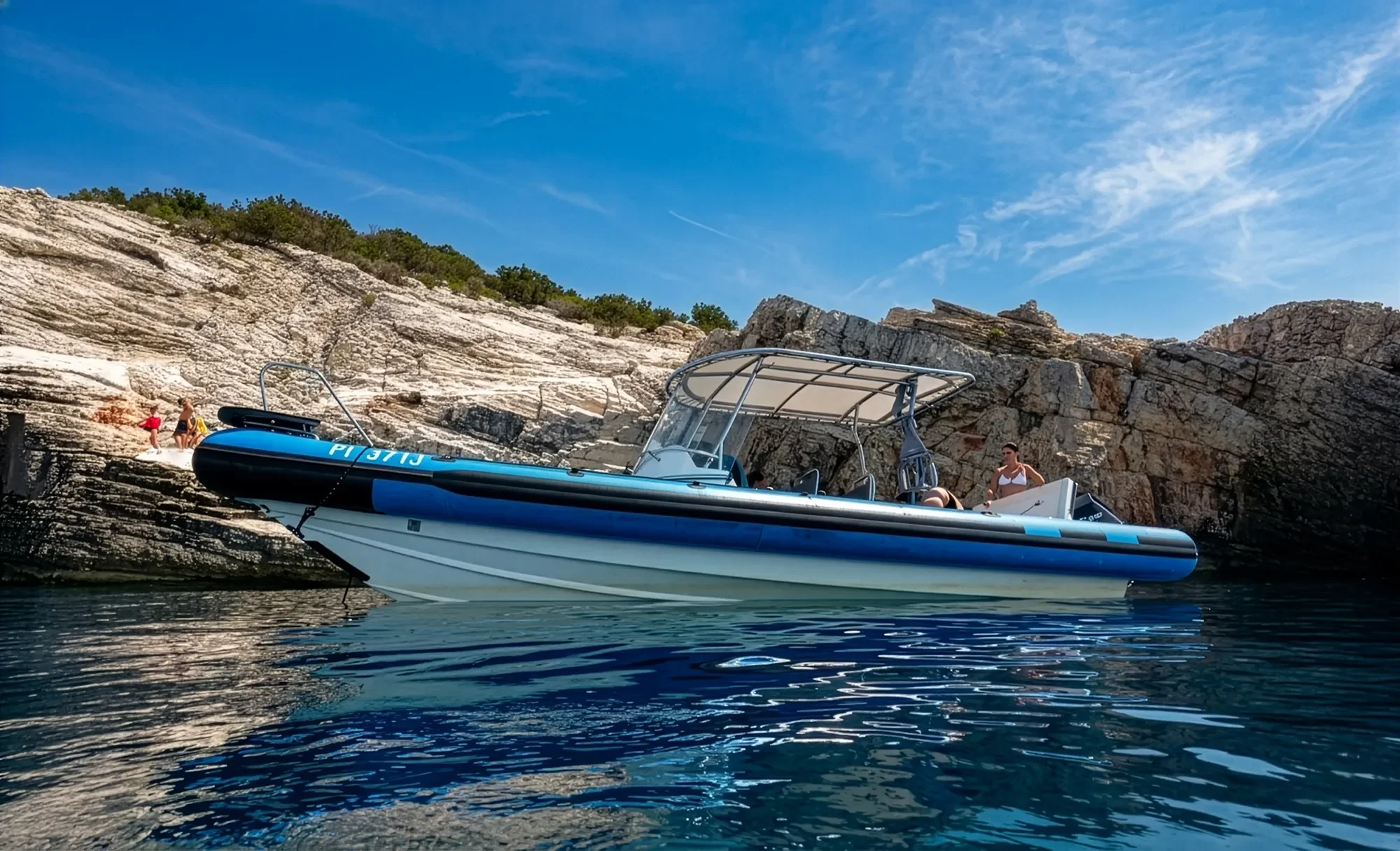 A blue and white inflatable boat with a canopy floating on calm water near rocky cliffs with a woman standing on the boat and three people on the rocky shore in the background under a clear blue sky.