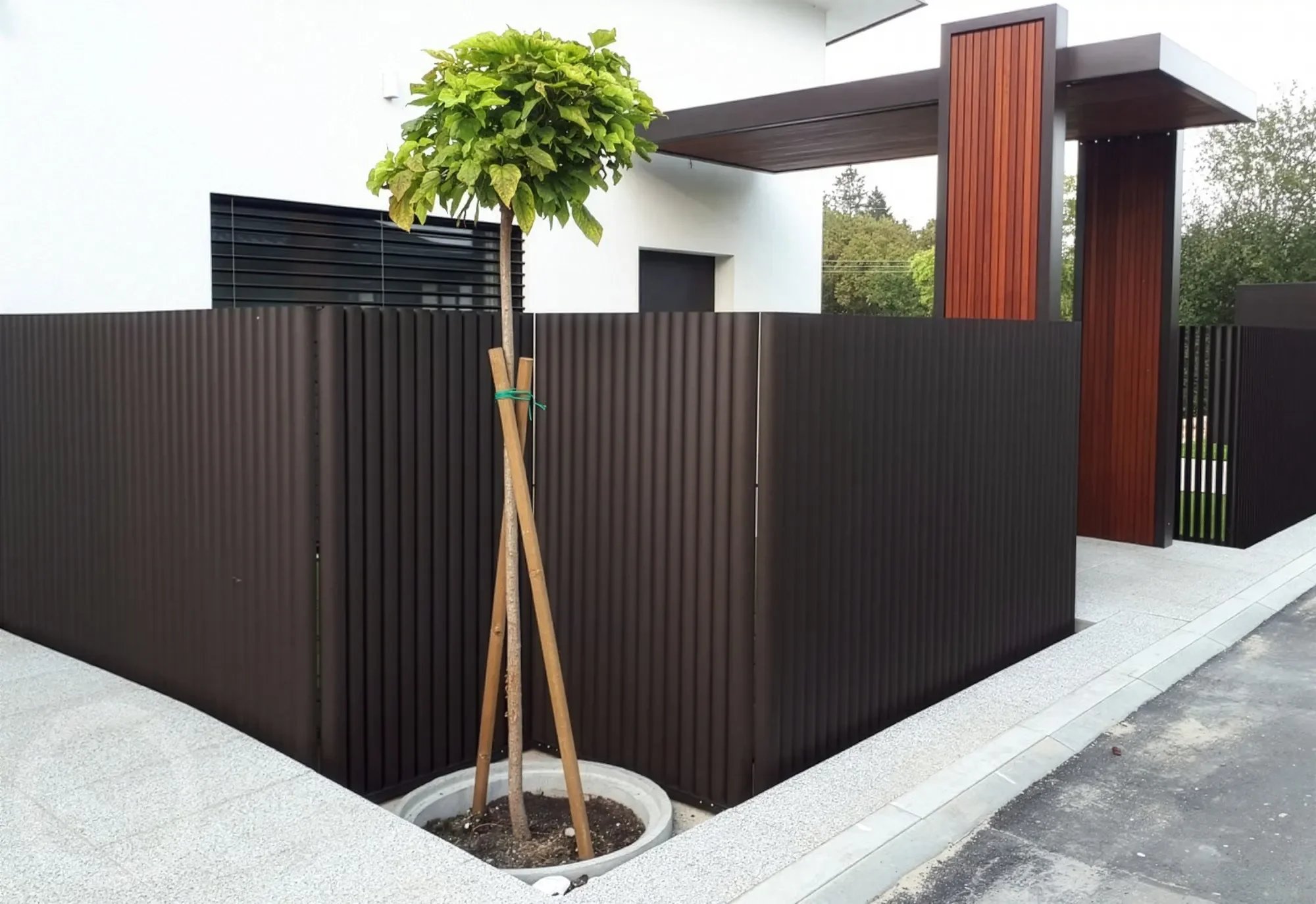 Modern house entrance with black corrugated metal fence, small tree in planter, and wooden accent panels over a white exterior wall.