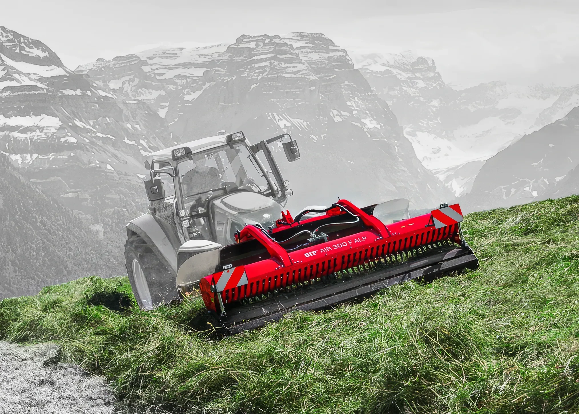A tractor with a red brush attachment cutting grass on a green hillside with a mountainous landscape in the background.
