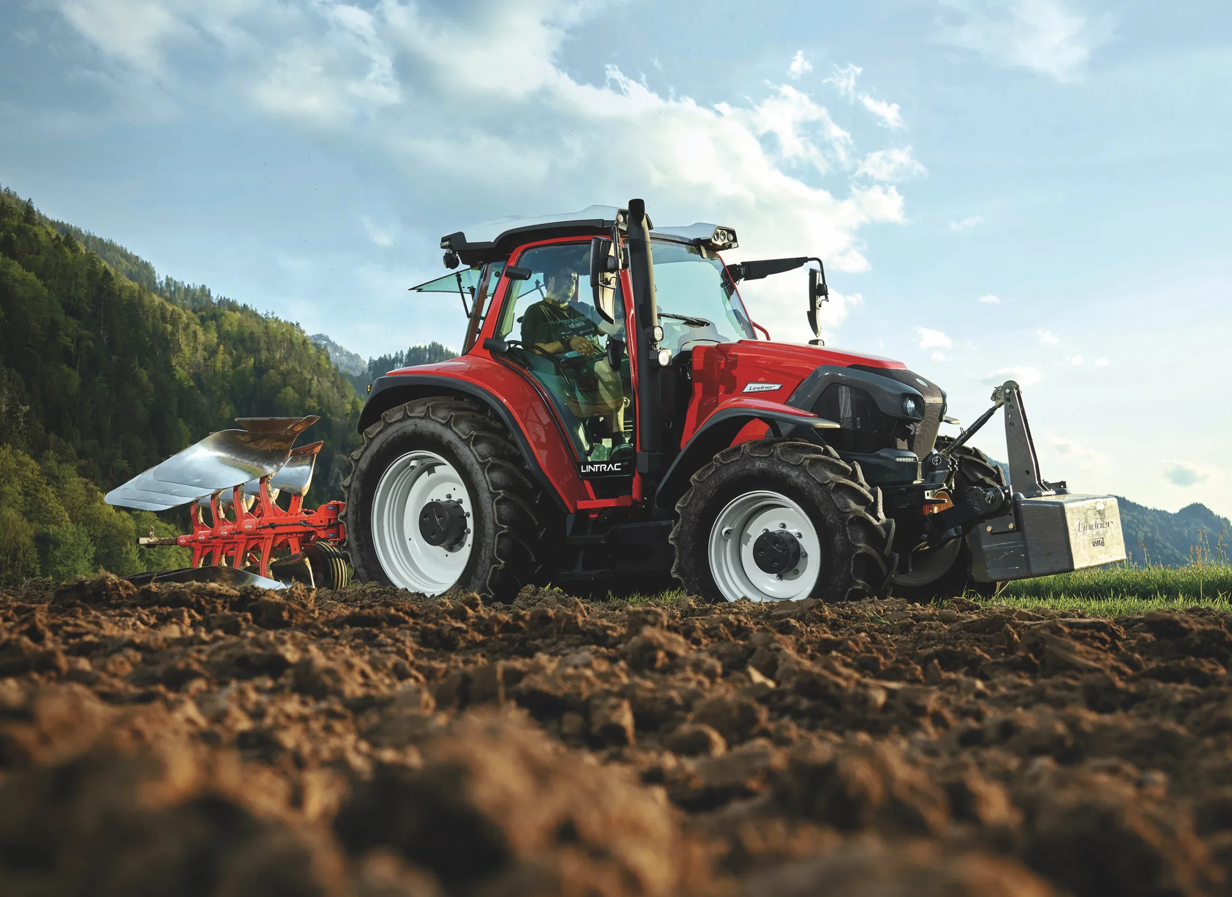 A red tractor with black tires working on a field with dirt, against a background of green hills and a partly cloudy sky.