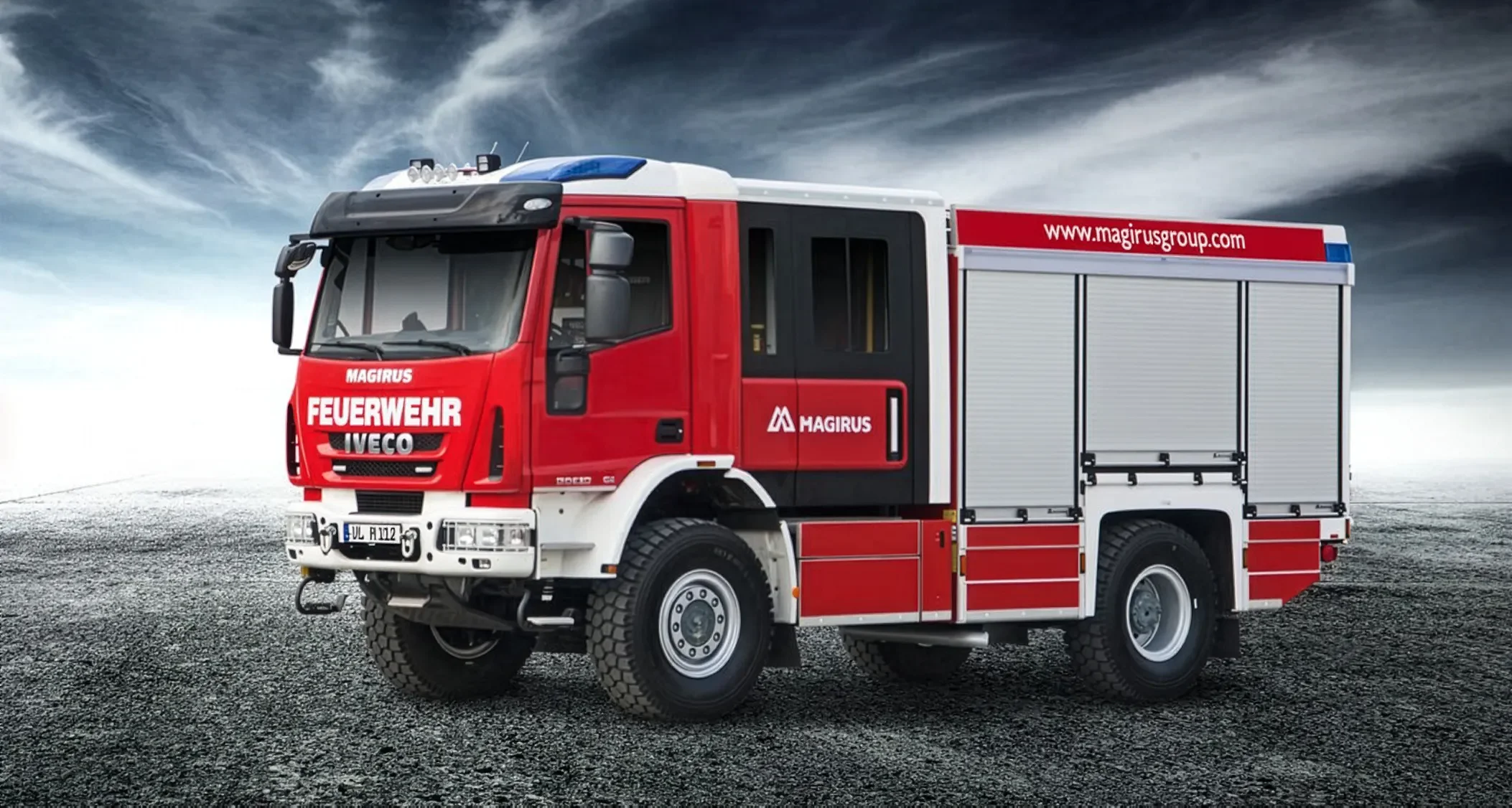 A red fire truck with white and black accents, marked with the words 'Feuerwehr' and 'Magirus', parked on a gravel surface under a cloudy sky.