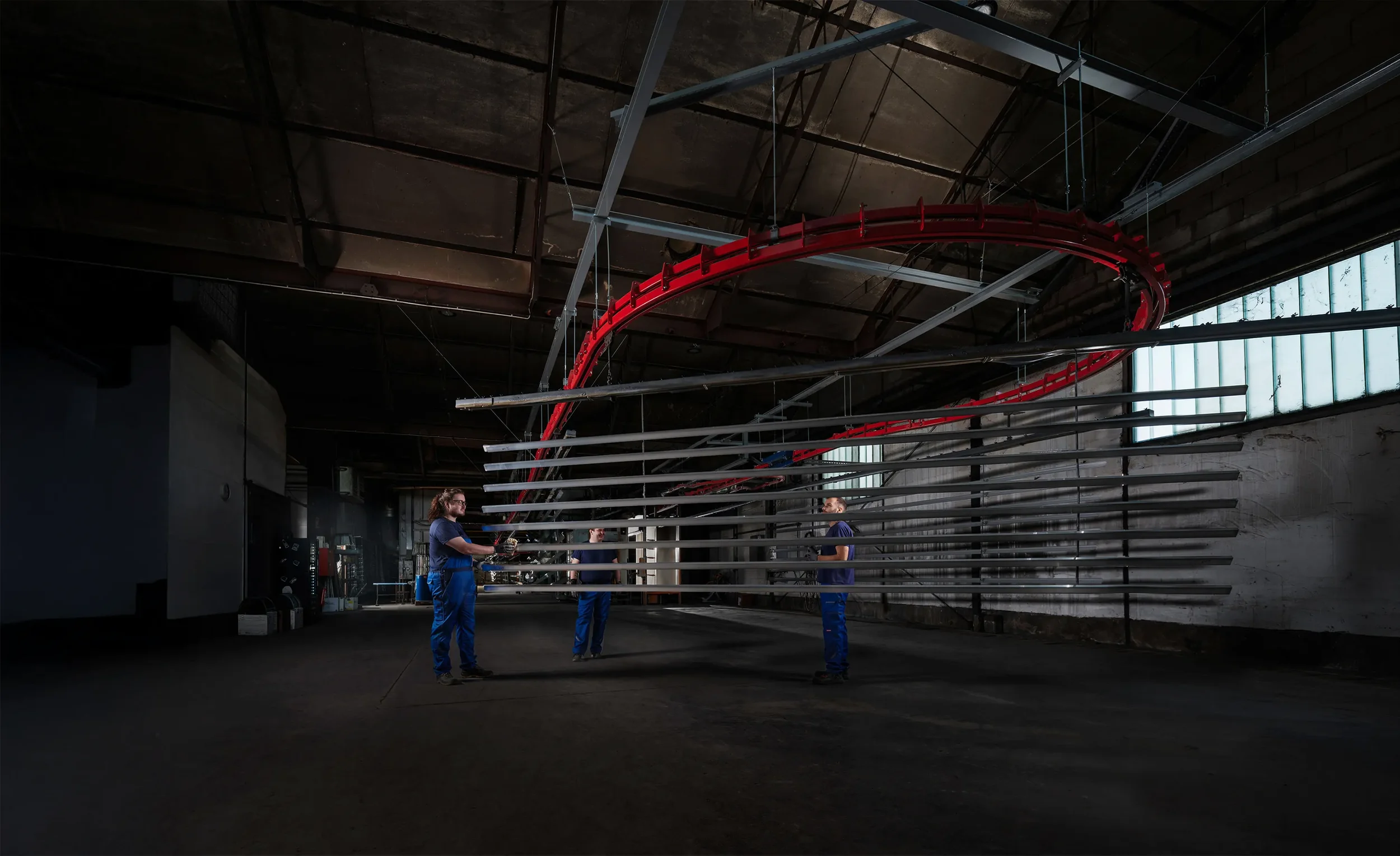 Three individuals in blue work uniforms operating a remote control inside an indoor facility with a large red roller coaster track overhead.