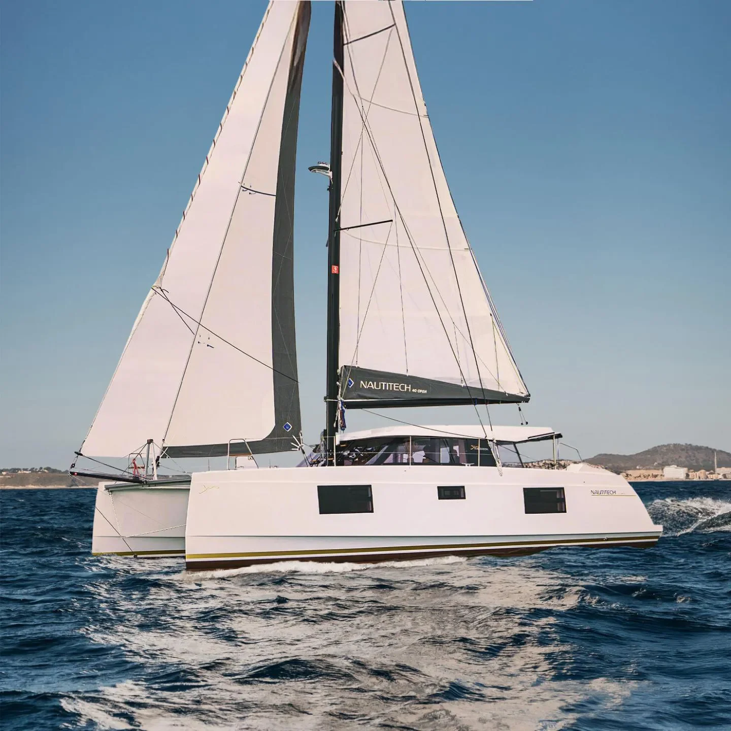A white sailing catamaran with black sails on open water, with land and a hill in the background.