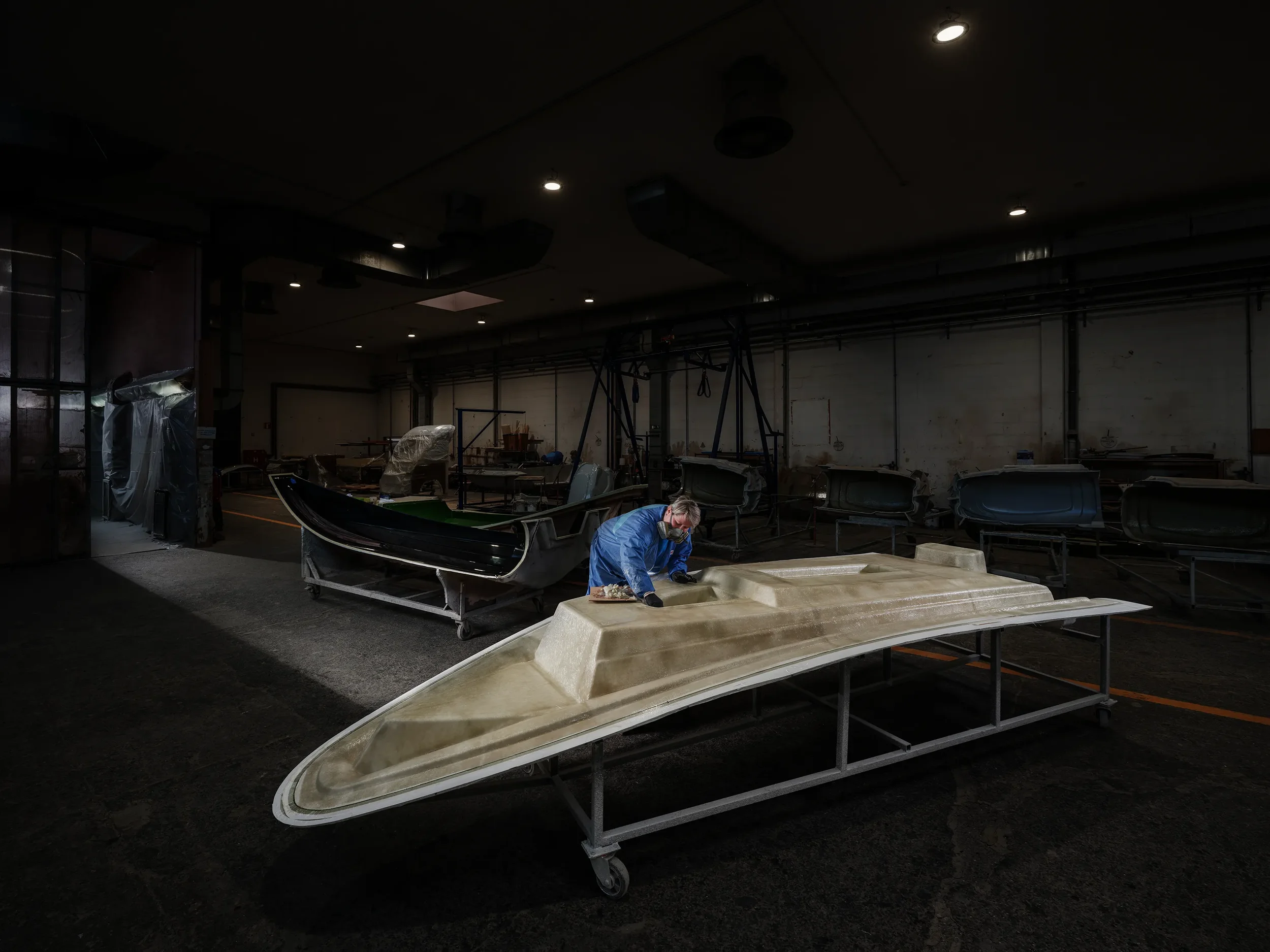 A worker wearing a mask and blue coveralls is working on a boat hull in a boat manufacturing or repair workshop. Multiple boats are visible in the background, some covered and some in assembly.