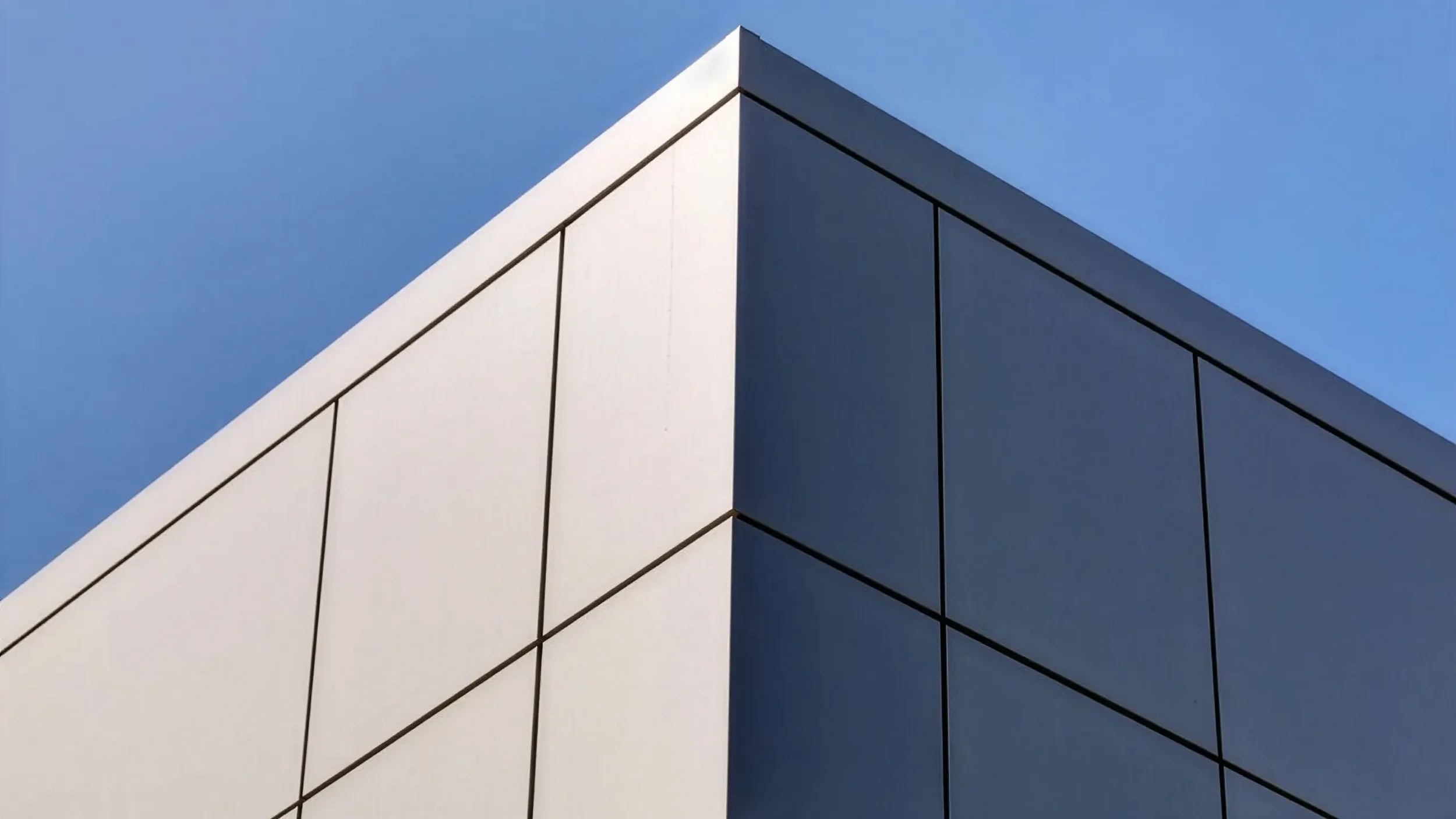 Close-up of a modern building's corner with white and dark gray panels against a blue sky.