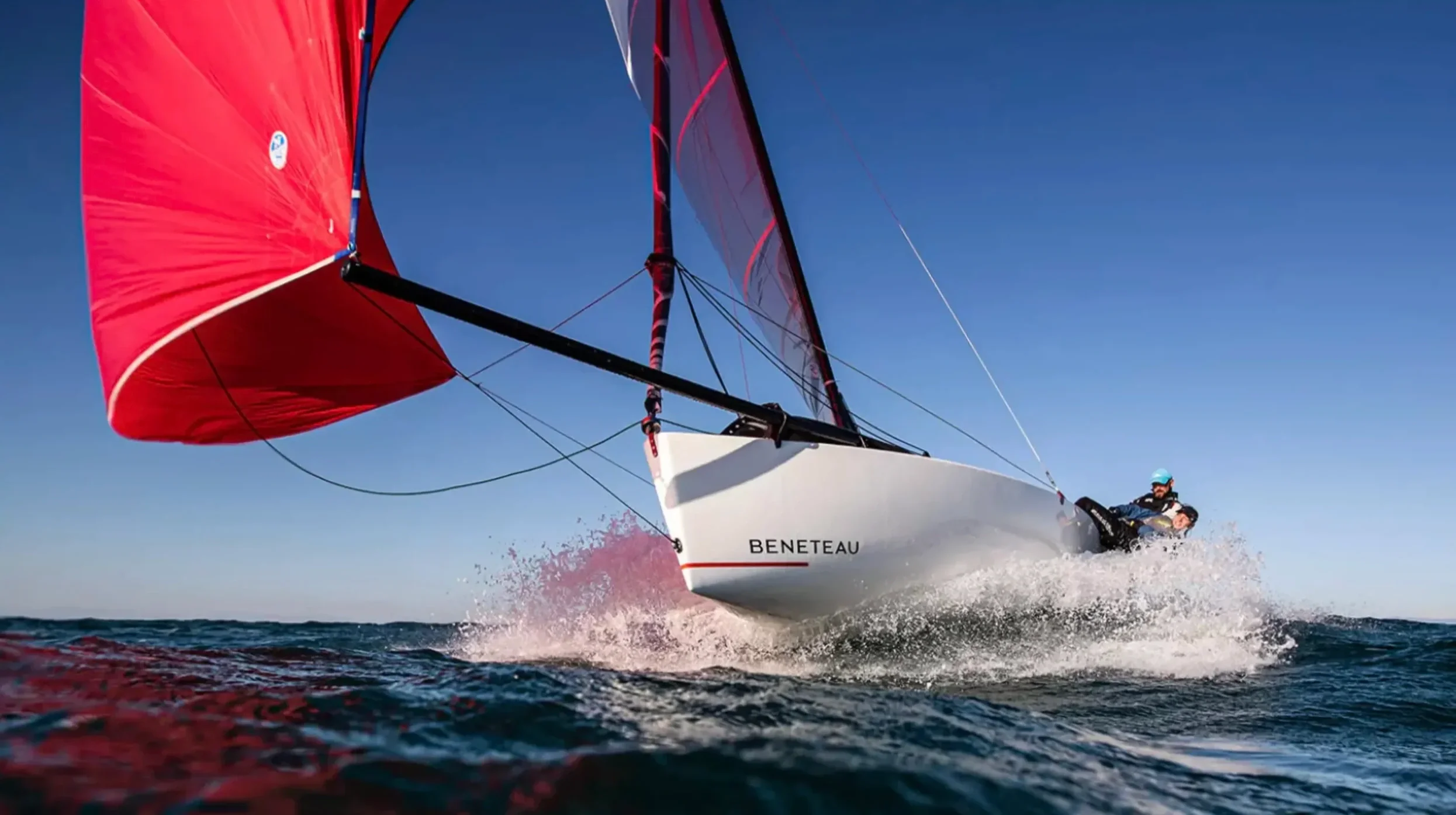 A sailboat named Beneteau with red sails moving quickly over the water, creating a splash, with a person in a blue helmet and dark clothing on board.