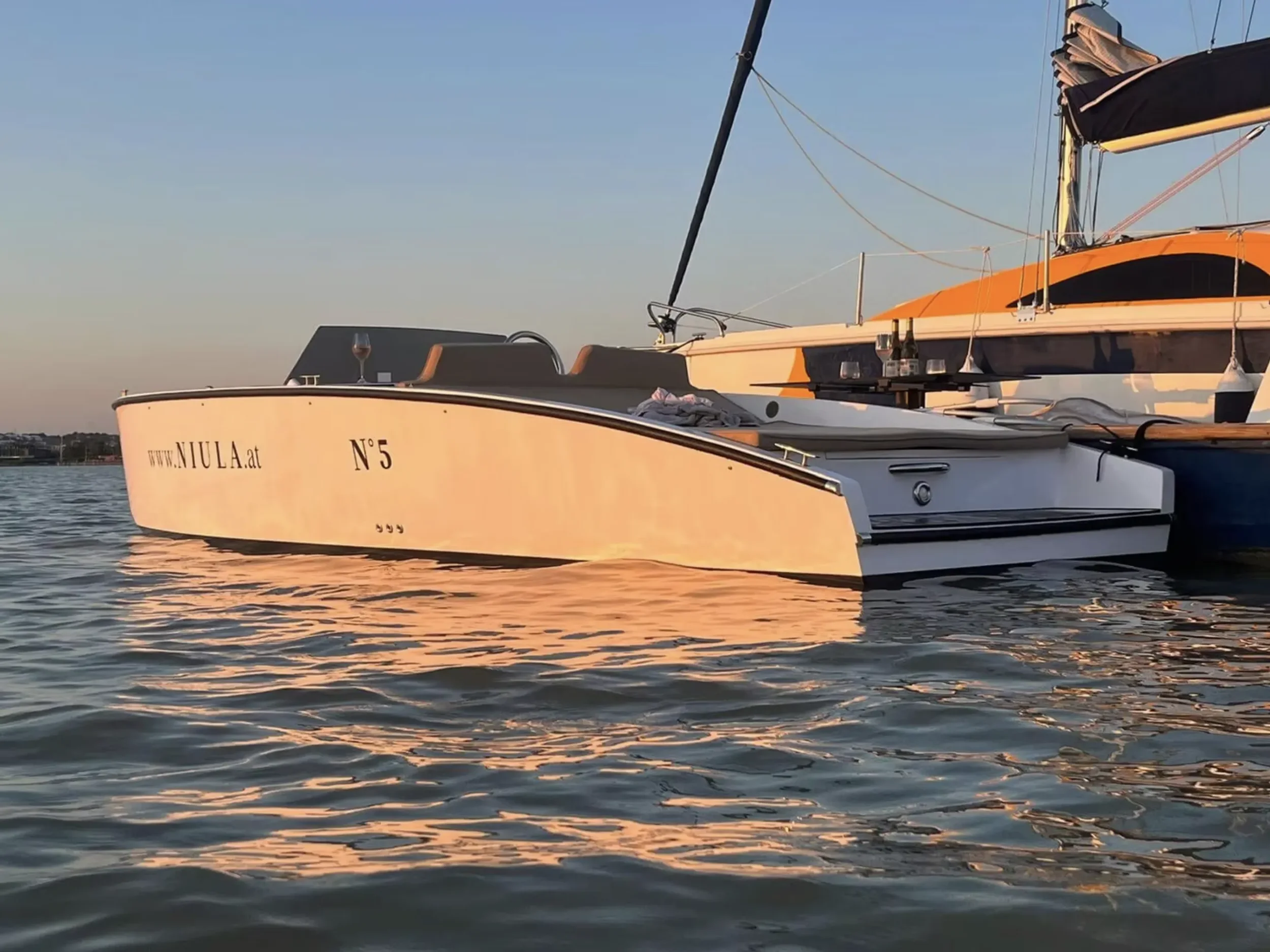 A white sailboat with the website and number 'www.NIULA.at N° 5' on its side, moored next to a larger orange and white yacht on calm water during sunset.