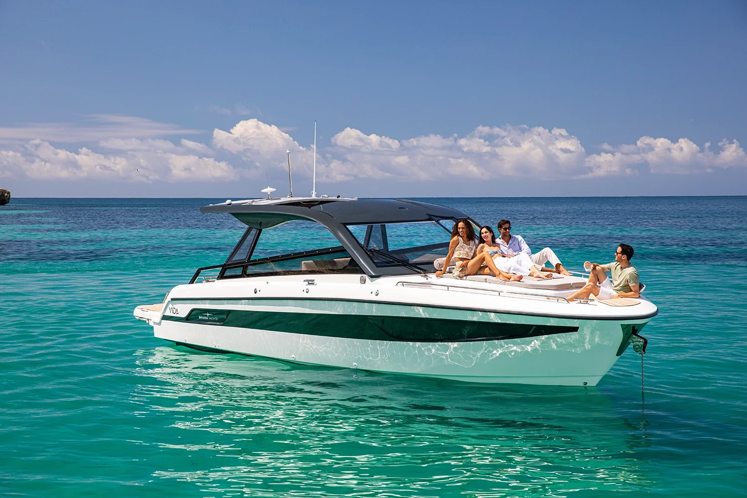 Four people relaxing on the deck of a white and green motorboat on turquoise water under a blue sky with clouds.